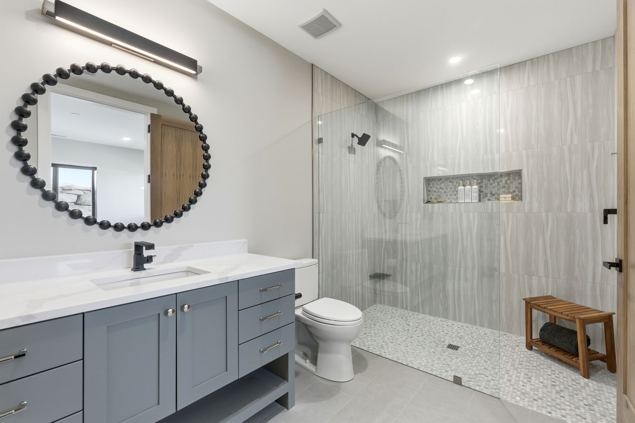 Modern bathroom featuring a single vanity with a white countertop and gray cabinetry, a round mirror with a beaded frame, and a walk-in shower with large format wall tiles and pebble-finish floor tiling