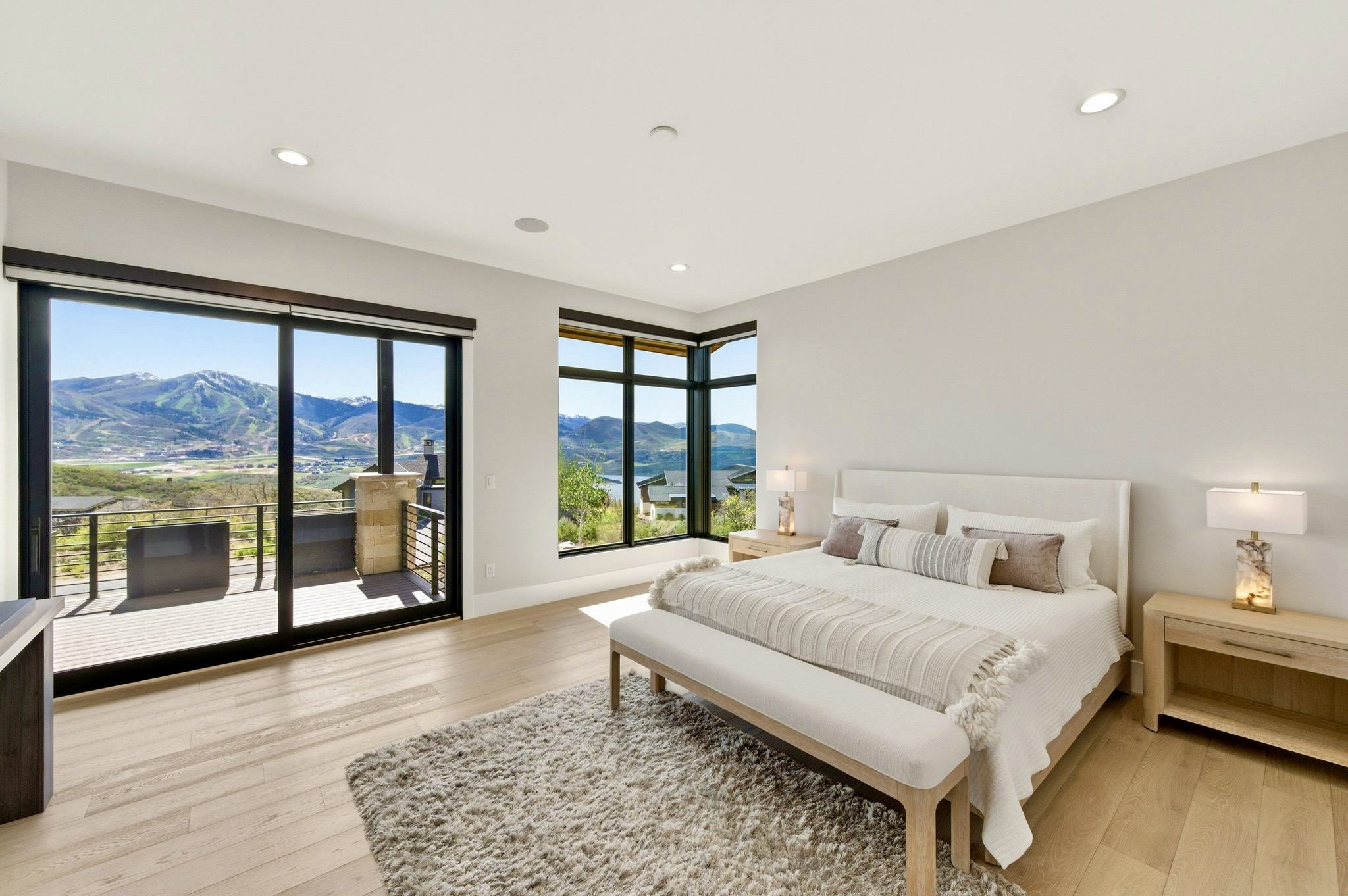 Expansive window walls showcasing mountain scenery, featuring light wood-finish flooring, recessed ceiling lighting, and a private outdoor balcony with metal railings and stone accents