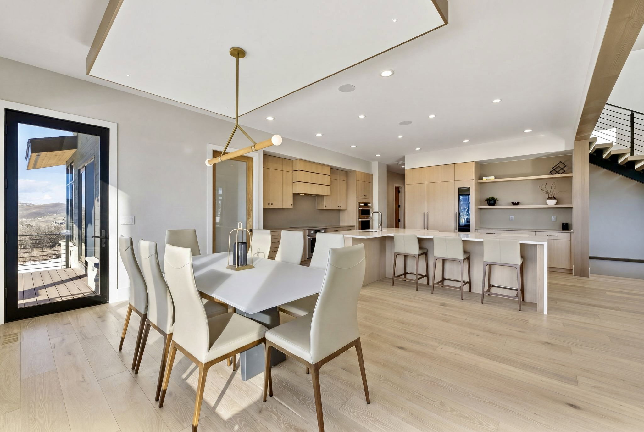 Open-concept living space featuring light wood-finish flooring, a contemporary dining area with a linear pendant light, an expansive kitchen island with bar seating, integrated modern appliances, and a glass-paneled door opening to an outdoor deck