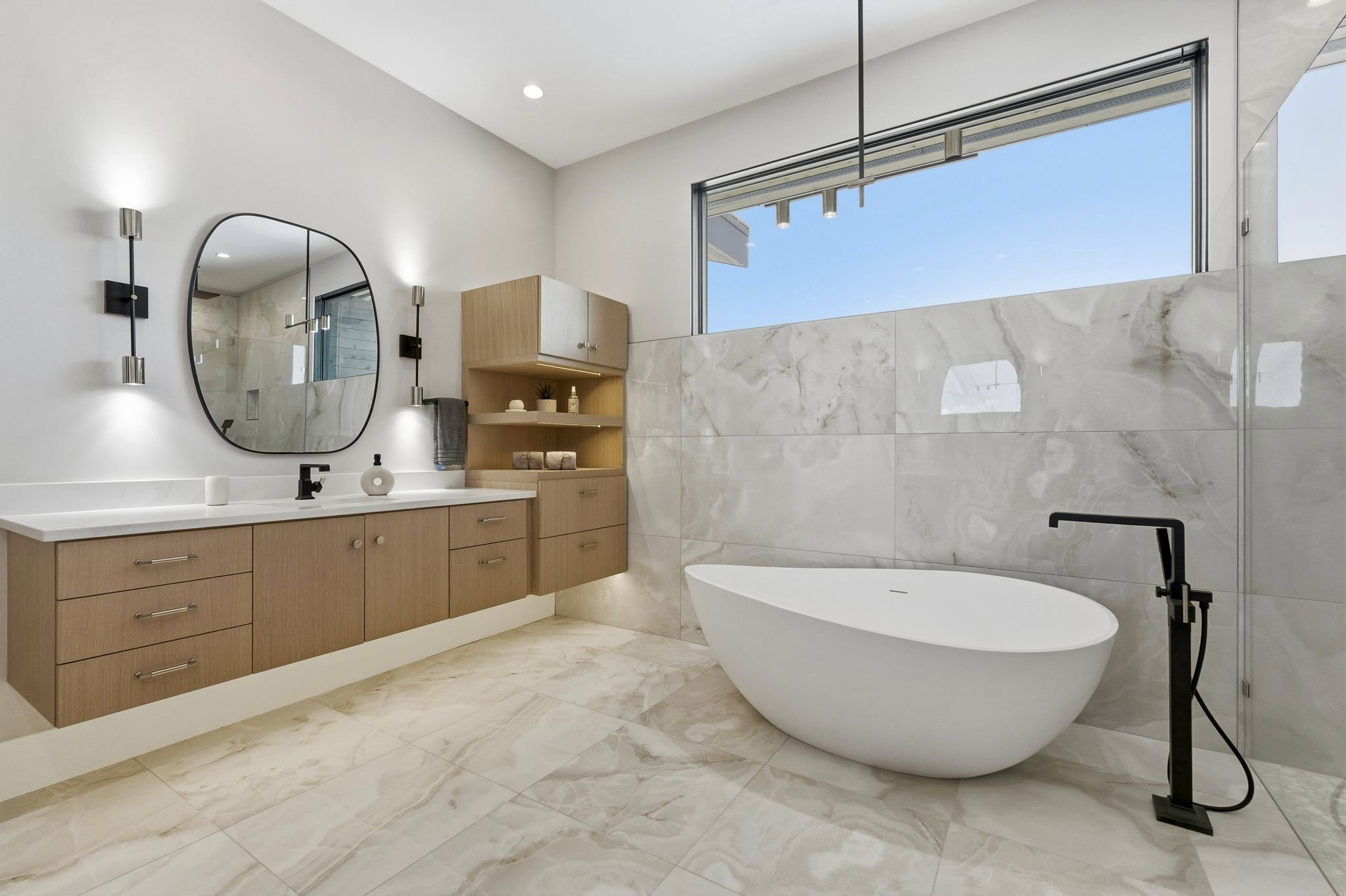 Contemporary bathroom featuring a freestanding soaking tub with a matte black floor-mounted tub filler, a floating vanity with a single basin, a rectangular window, large format tile flooring, and wall-mounted sconces