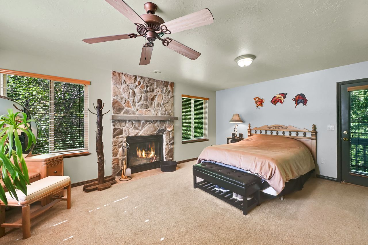 Stone-surround fireplace with a hearth and mantel, complemented by two windows with wood blinds, a ceiling fan, and a glass-paneled door