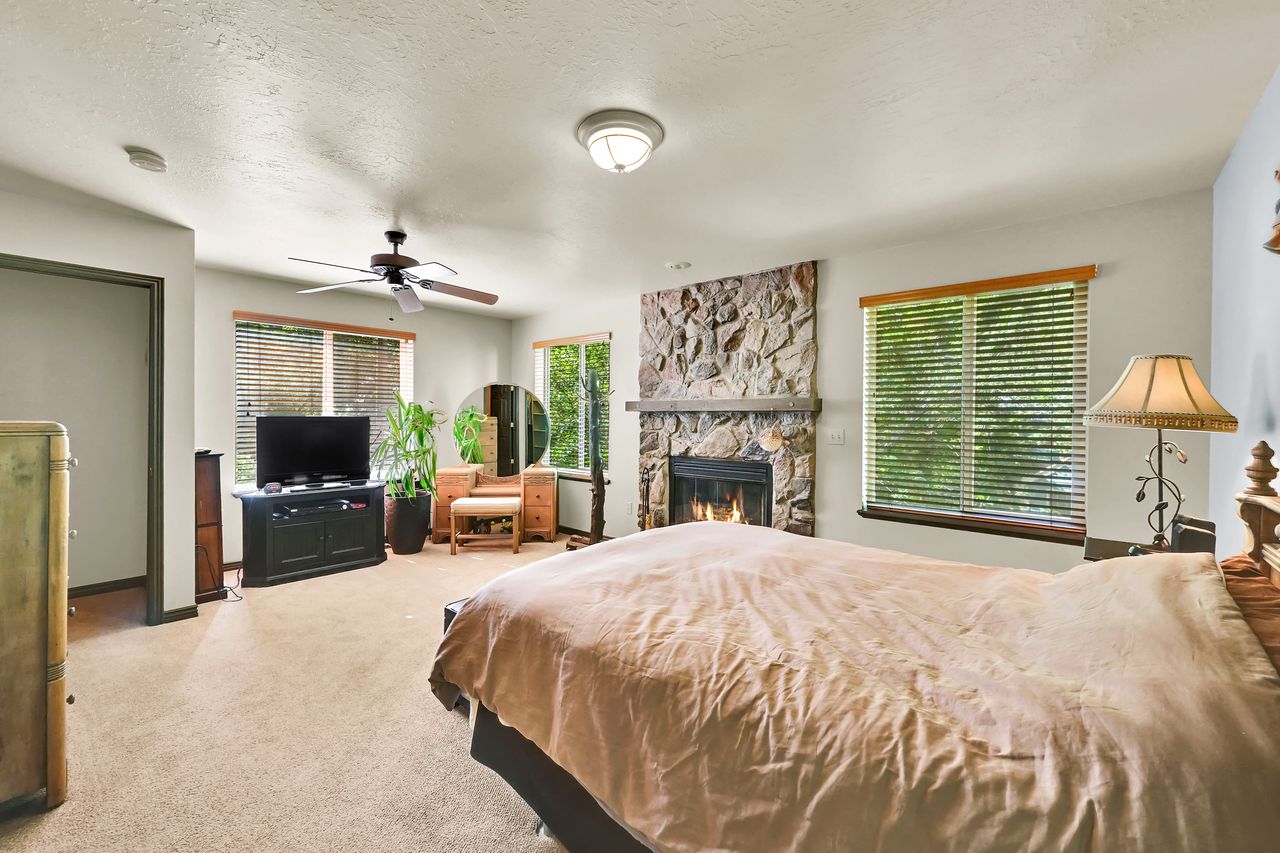 Spacious room featuring a stone-clad fireplace, ceiling fan, carpet flooring, and multiple windows with blinds
