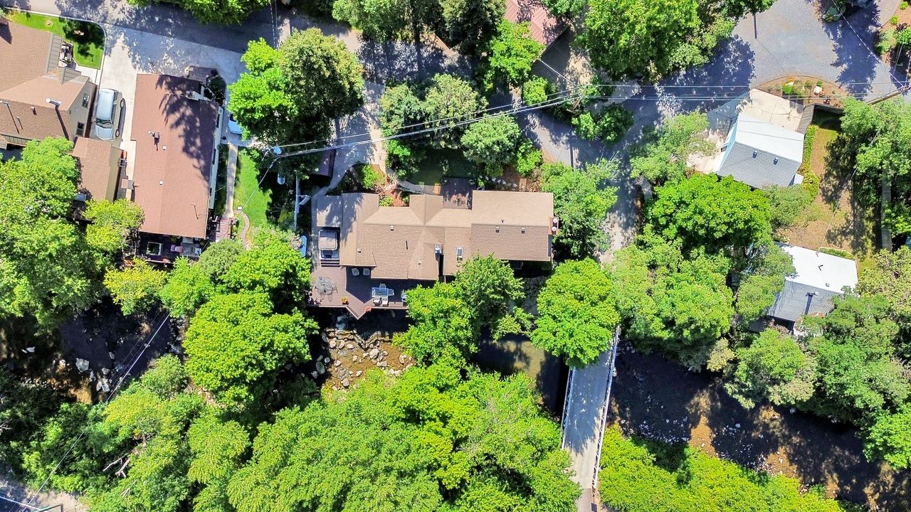 Aerial perspective showcasing a property surrounded by mature trees, featuring a brown shingle roof and a multi-level deck overlooking a natural creek