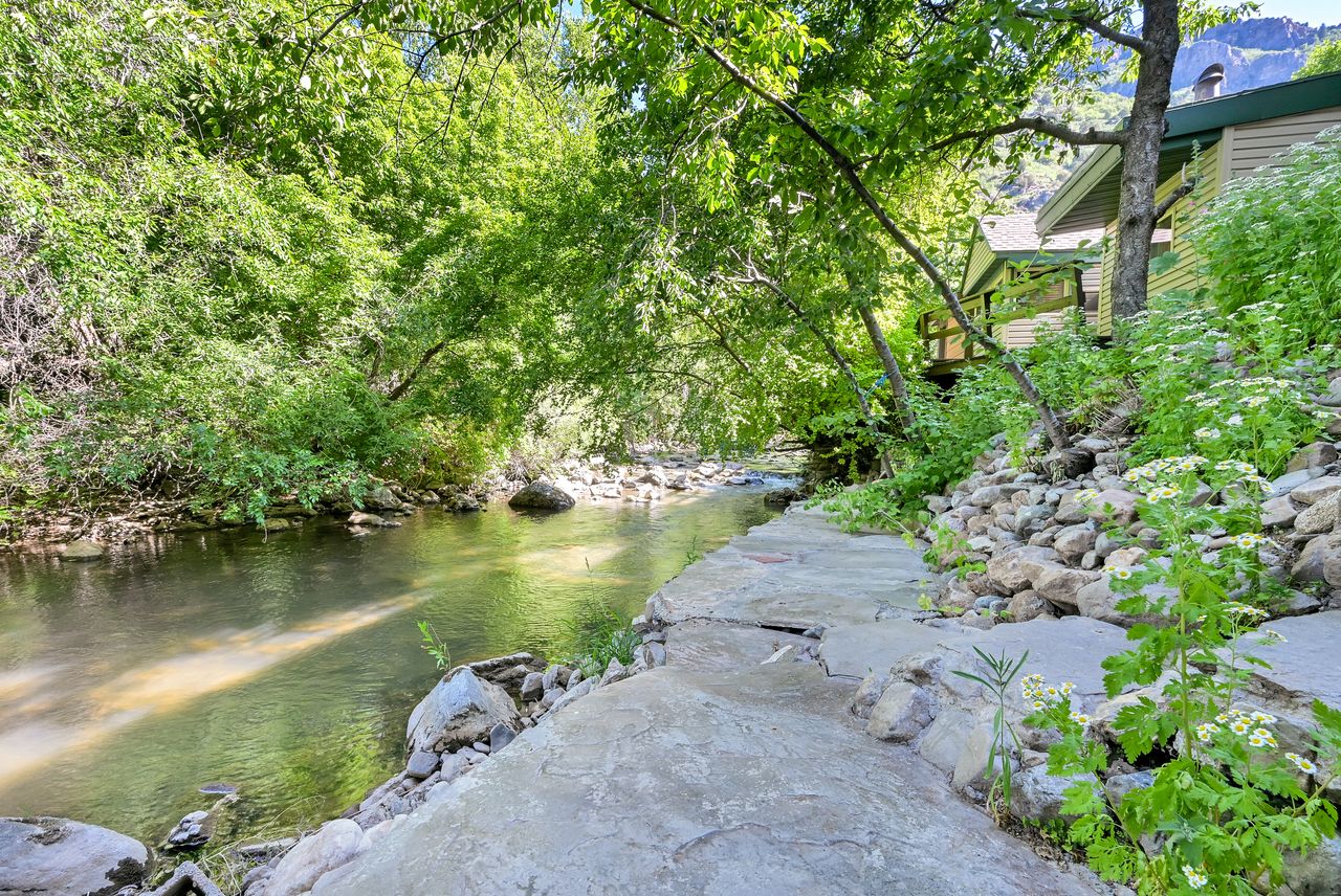 Riverfront property featuring a stone path, lush surrounding trees, and a distant house with a balcony