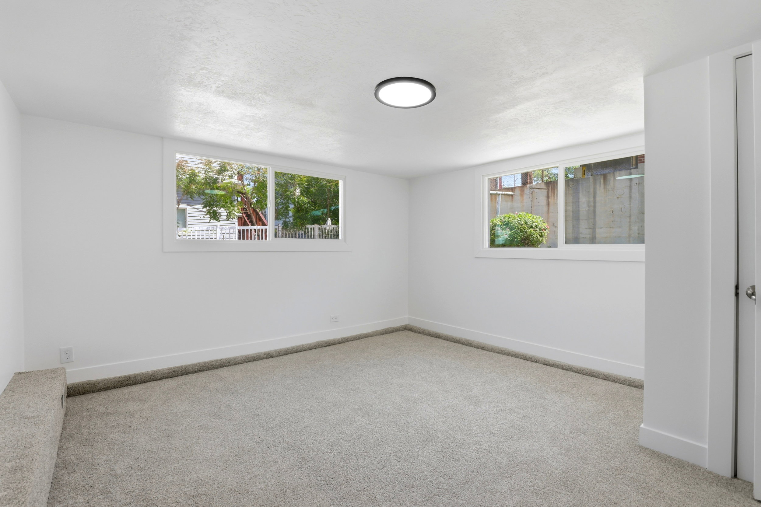 Spacious room featuring light gray carpeting, crisp white walls, multiple windows, a modern flush-mount ceiling light, and white baseboards