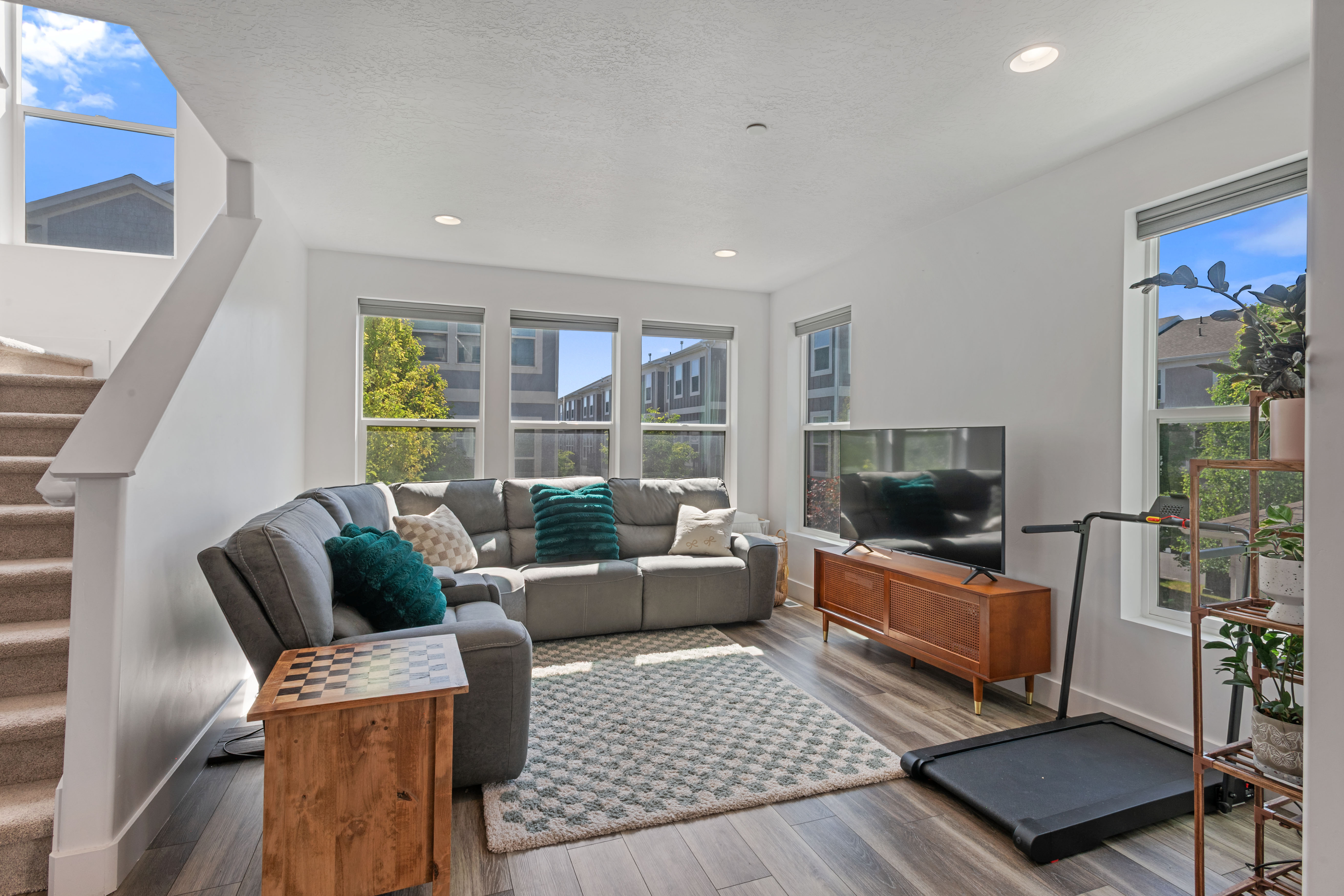Bright living area featuring wood-finish flooring, recessed lighting, multiple large windows, and a carpeted staircase with a white railing