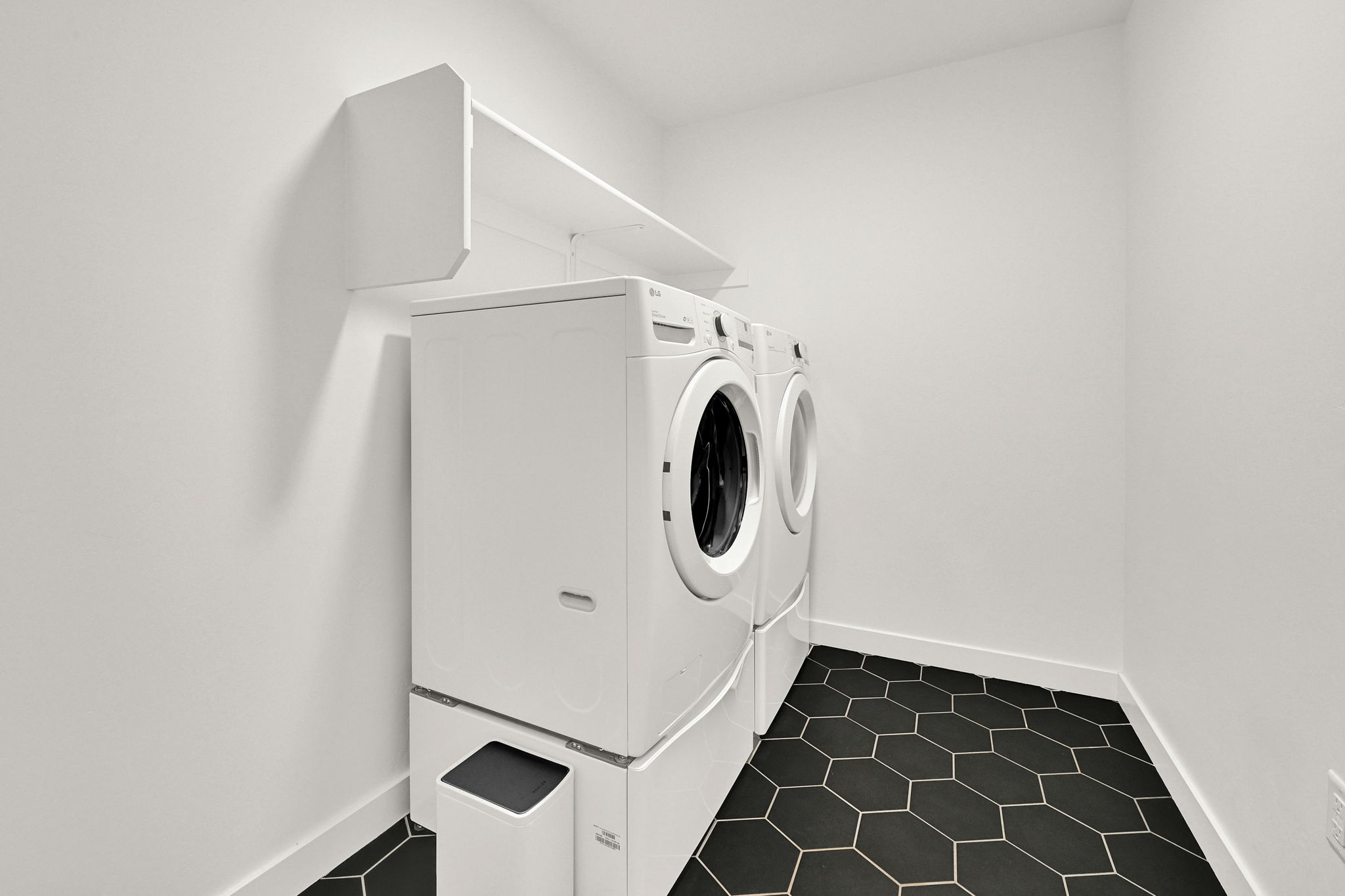 Dedicated laundry area featuring a white front-load washer and dryer set, elevated on pedestals, with overhead shelving and a hexagonal tile floor