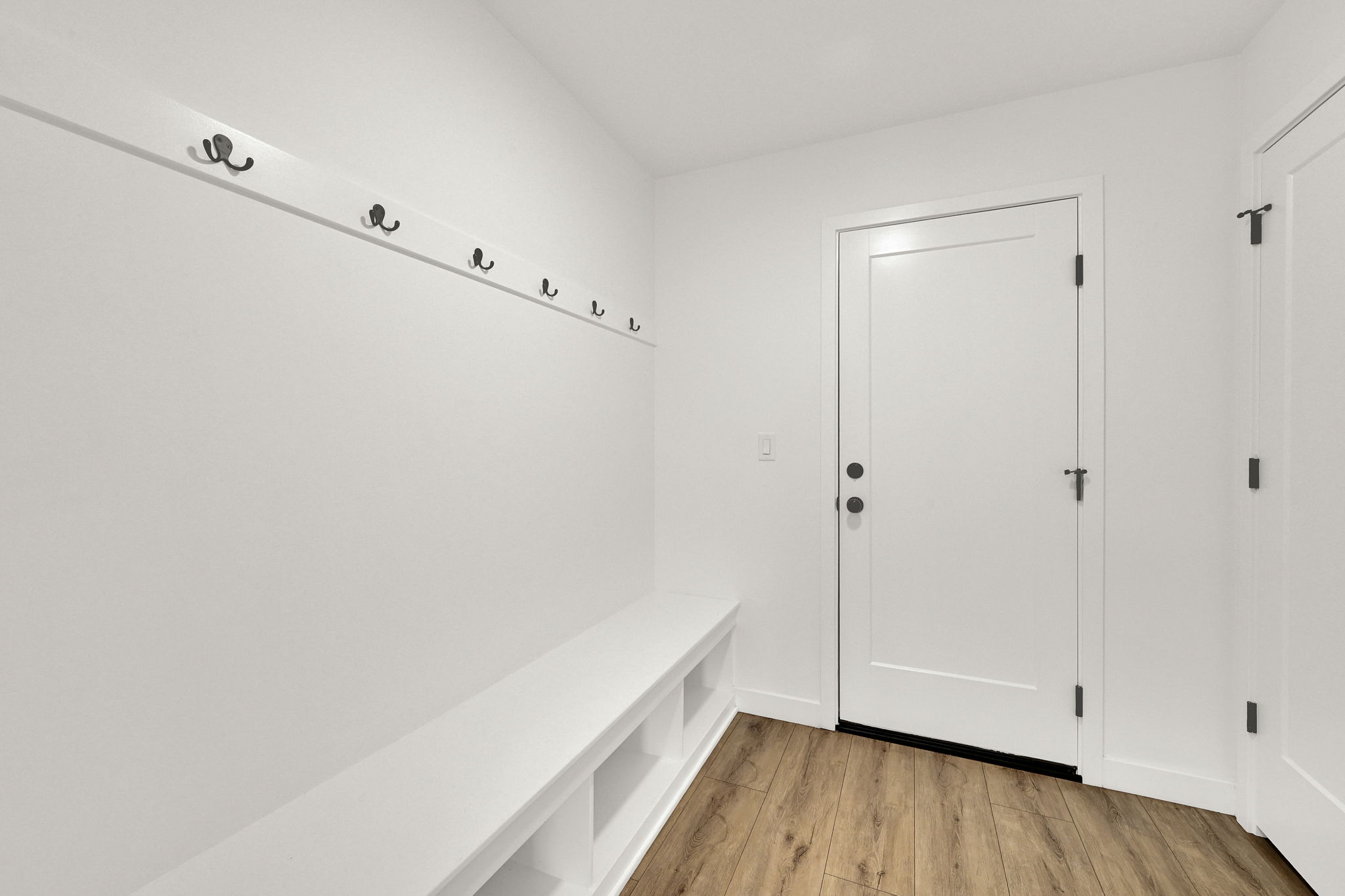 Entryway featuring white built-in bench seating with cubbies, a white wall-mounted coat rack with black hooks, a white door with black hardware, and wood-finish flooring