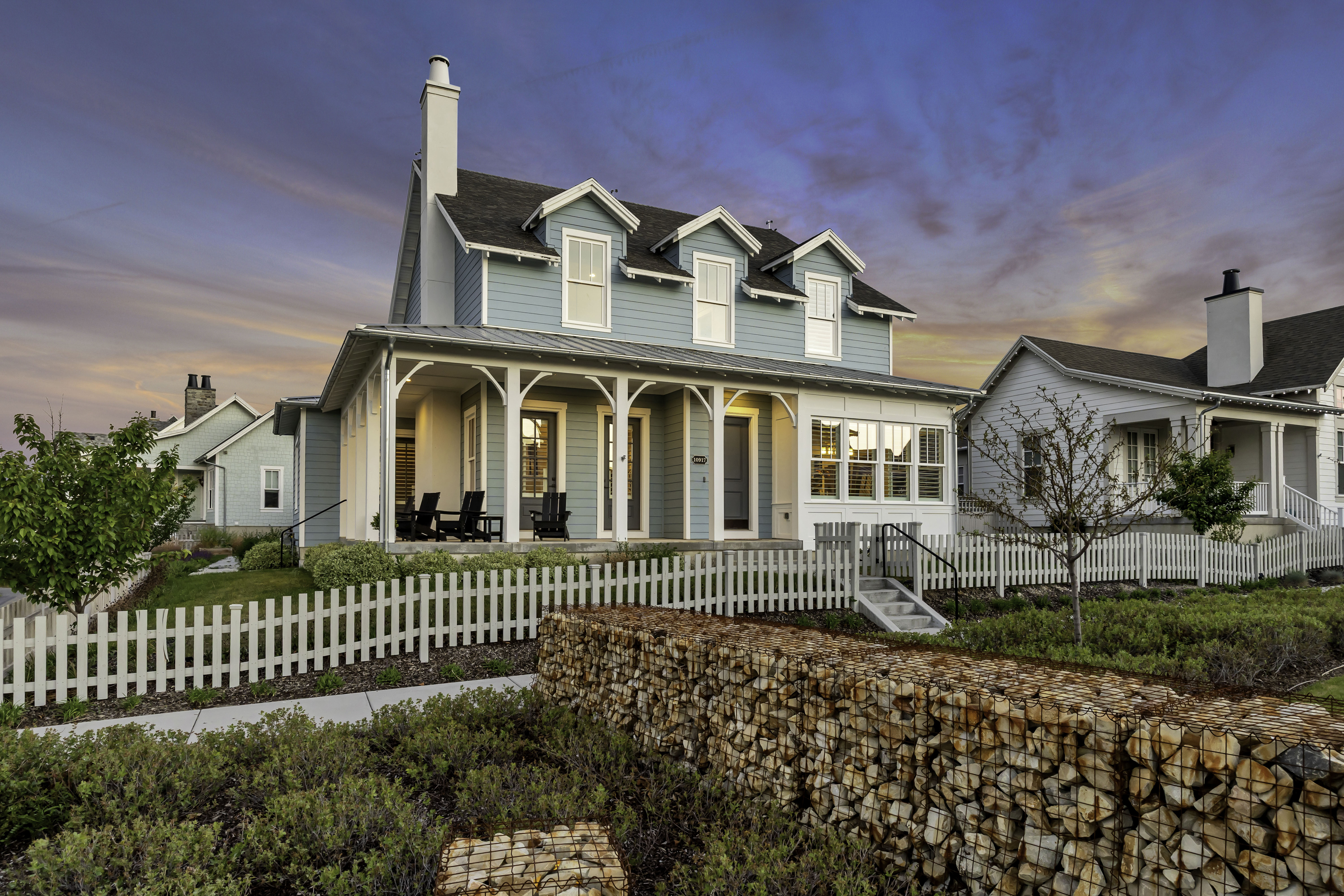 Blue siding exterior with white trim, a covered front porch, multiple dormer windows, a prominent chimney, and a white picket fence