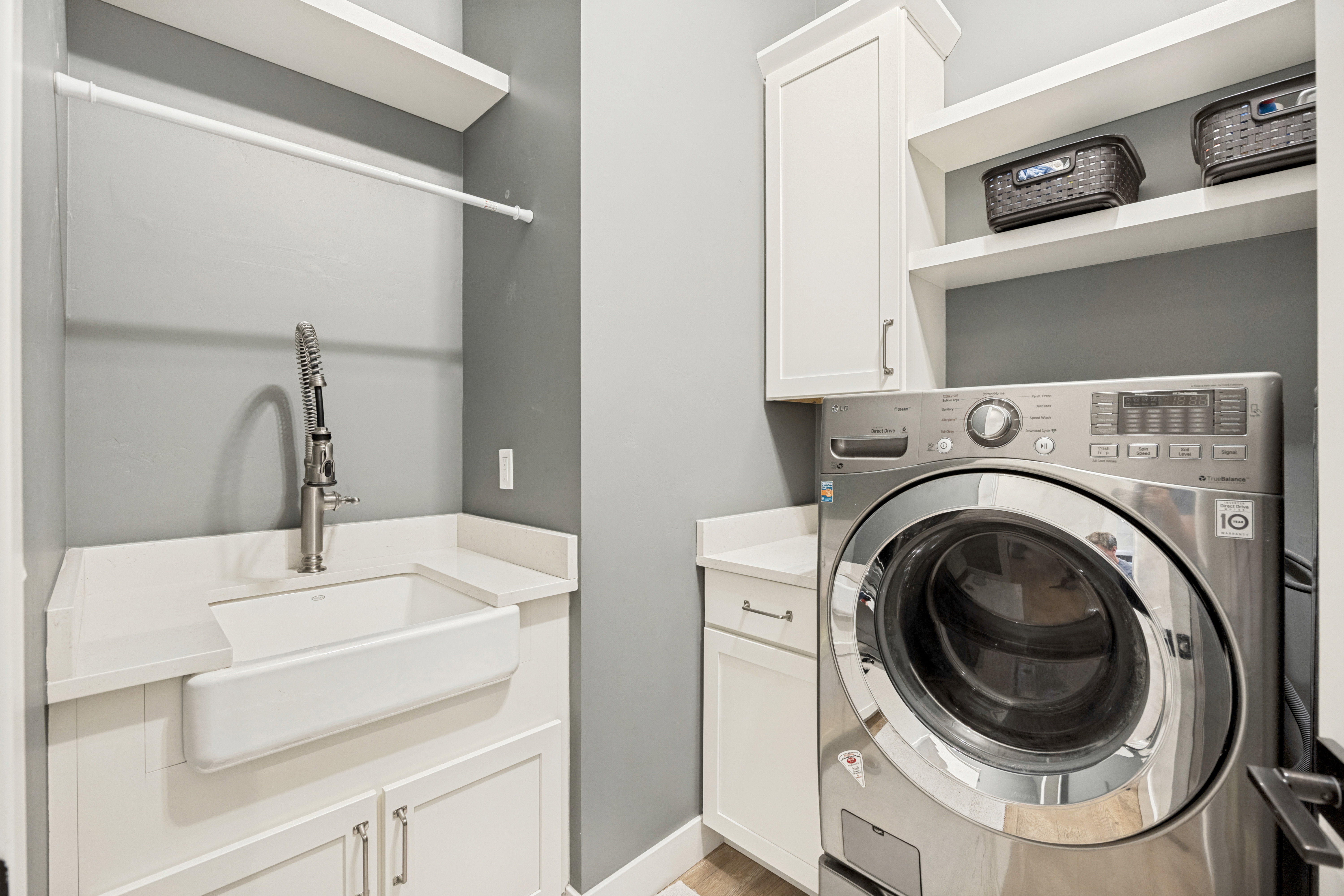 Dedicated laundry space featuring a farmhouse sink with a pull-down faucet, white cabinetry, solid surface countertops, a built-in hanging rod, and open shelving