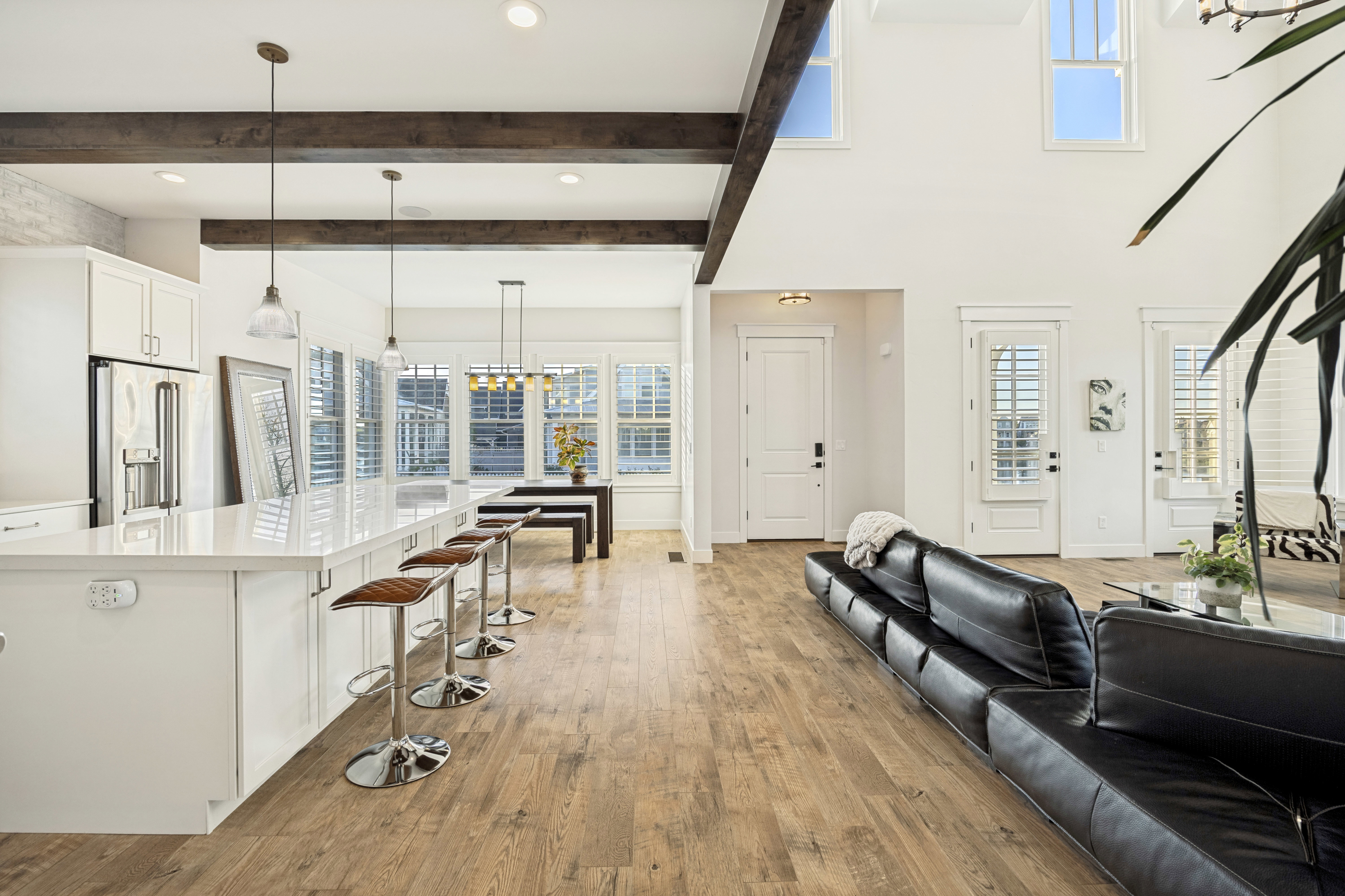 Open-concept living area featuring wood-finish flooring, exposed ceiling beams, a kitchen island with seating, white cabinetry, and multiple windows with plantation shutters