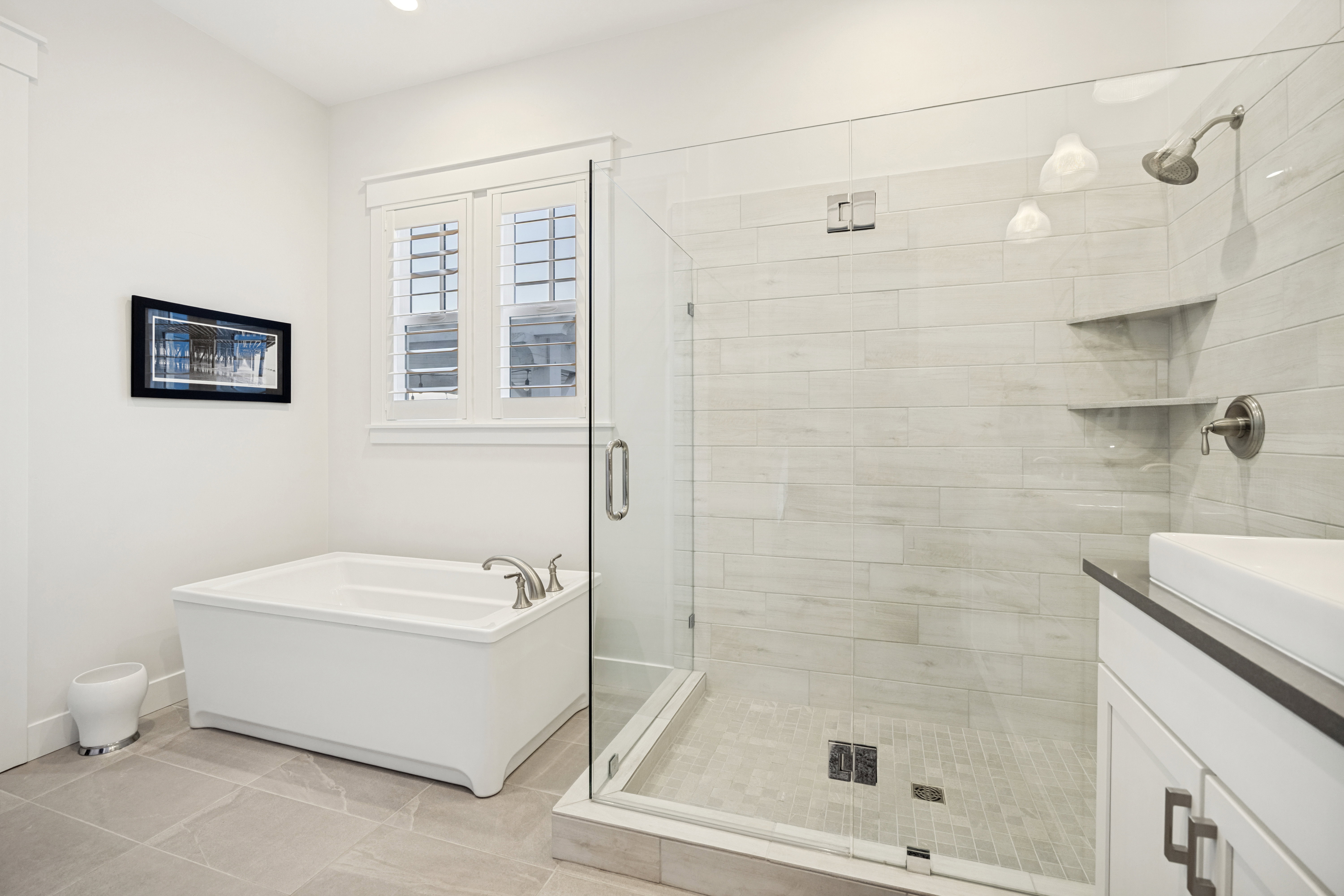Bathroom featuring a glass-enclosed shower with horizontal tile walls and built-in shelving, a separate soaking tub with brushed nickel fixtures, and a white vanity with a dark countertop and undermount sink
