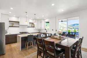 Open-concept kitchen and dining area featuring light wood-finish flooring, a kitchen island with seating, a gas range with a modern vent hood, white upper cabinetry, and dark wood lower cabinetry