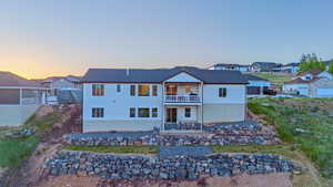 Two-story residence featuring a white stucco exterior with a dark shingle roof, upper-level balcony with white railings, a lower-level patio, and tiered rock retaining walls