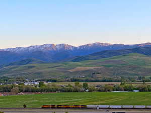 Expansive landscape featuring snow-capped mountains, verdant rolling hills, a distant town, and a railway track with a passing train