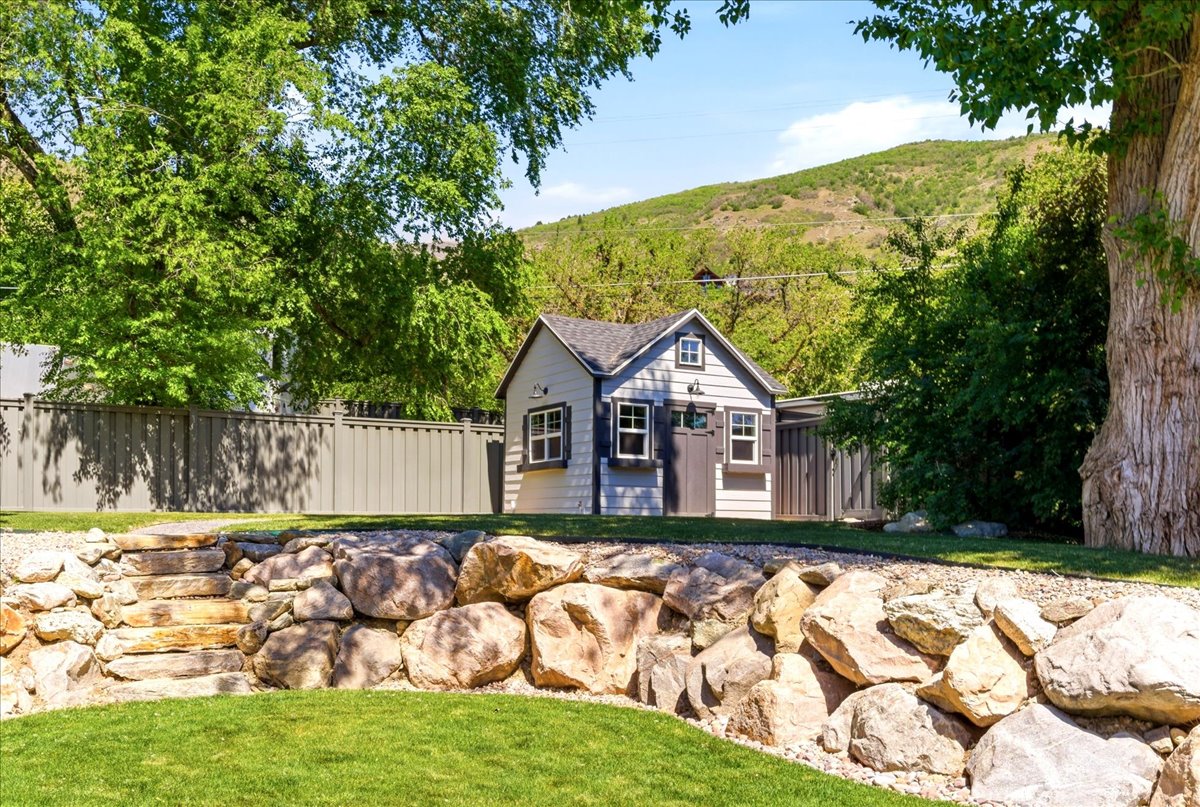 Landscaped backyard featuring a tiered rock retaining wall with stone steps, a manicured lawn, and a custom storage shed with board and batten siding