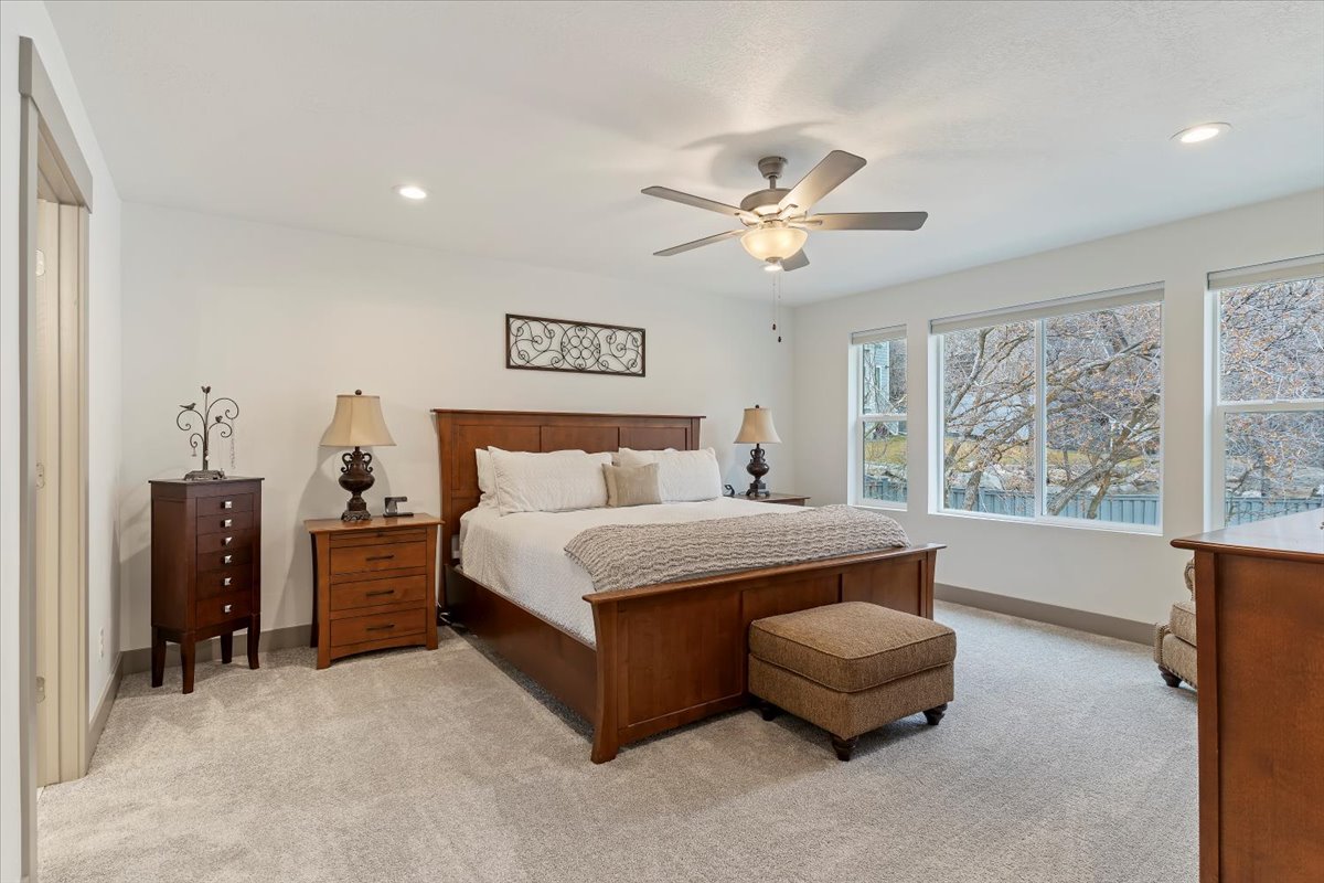 Spacious bedroom featuring light-toned carpeting, recessed lighting, a ceiling fan with integrated lighting, and multiple large windows providing abundant natural light