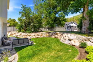 Expansive backyard featuring a manicured lawn, a rock retaining wall, a dedicated patio area, mature trees, and a detached shed