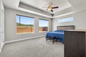 Spacious bedroom featuring light gray walls, plush carpet flooring, a tiered tray ceiling with a fan, and large windows providing ample natural light