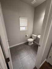 Bathroom featuring gray wall paint, a white window with blinds, a white toilet, and gray tile flooring