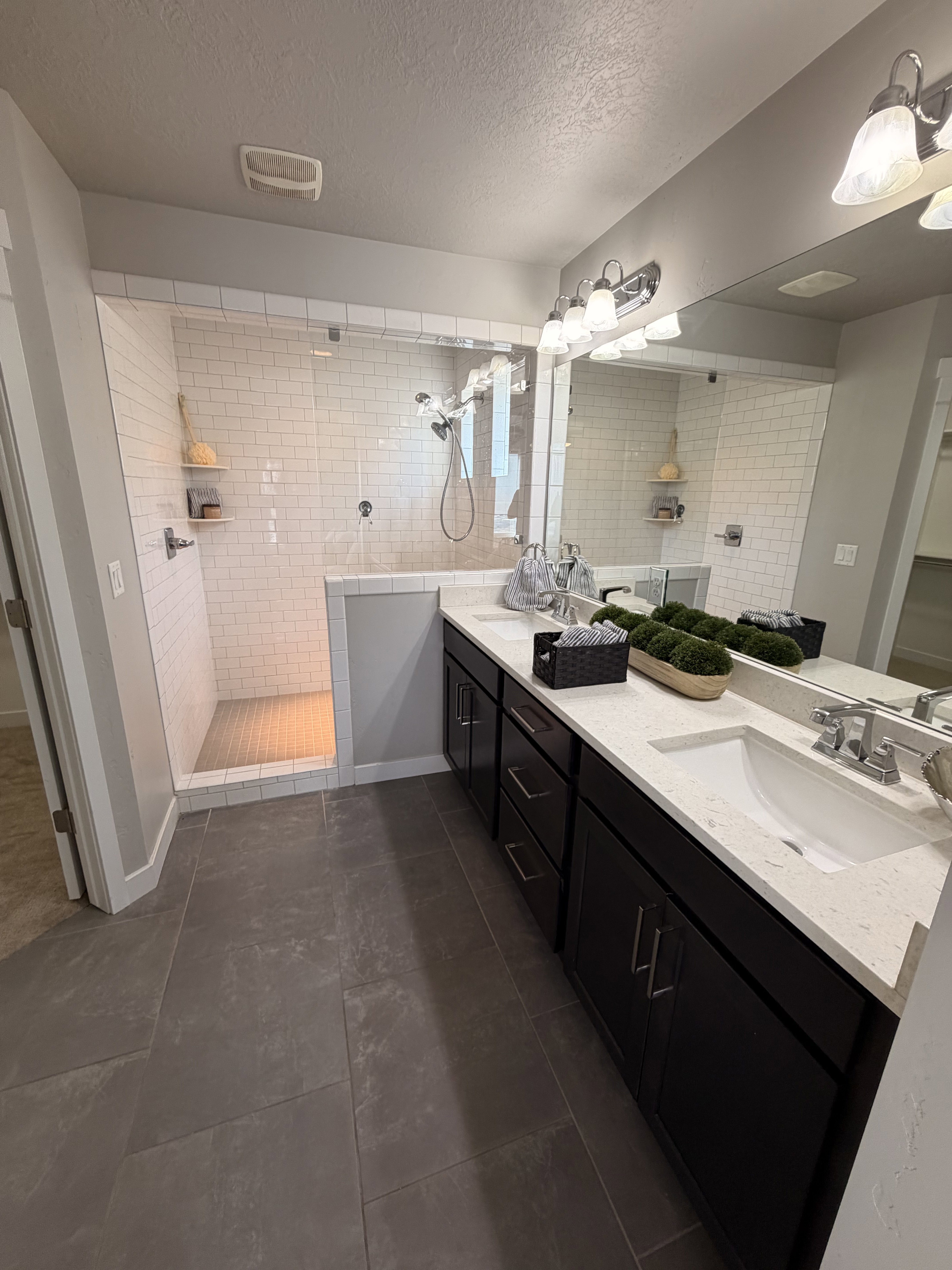 Spacious bathroom featuring a double vanity with dark cabinetry and light-toned countertops, a large mirror, and a walk-in shower with subway tile surround and glass enclosure