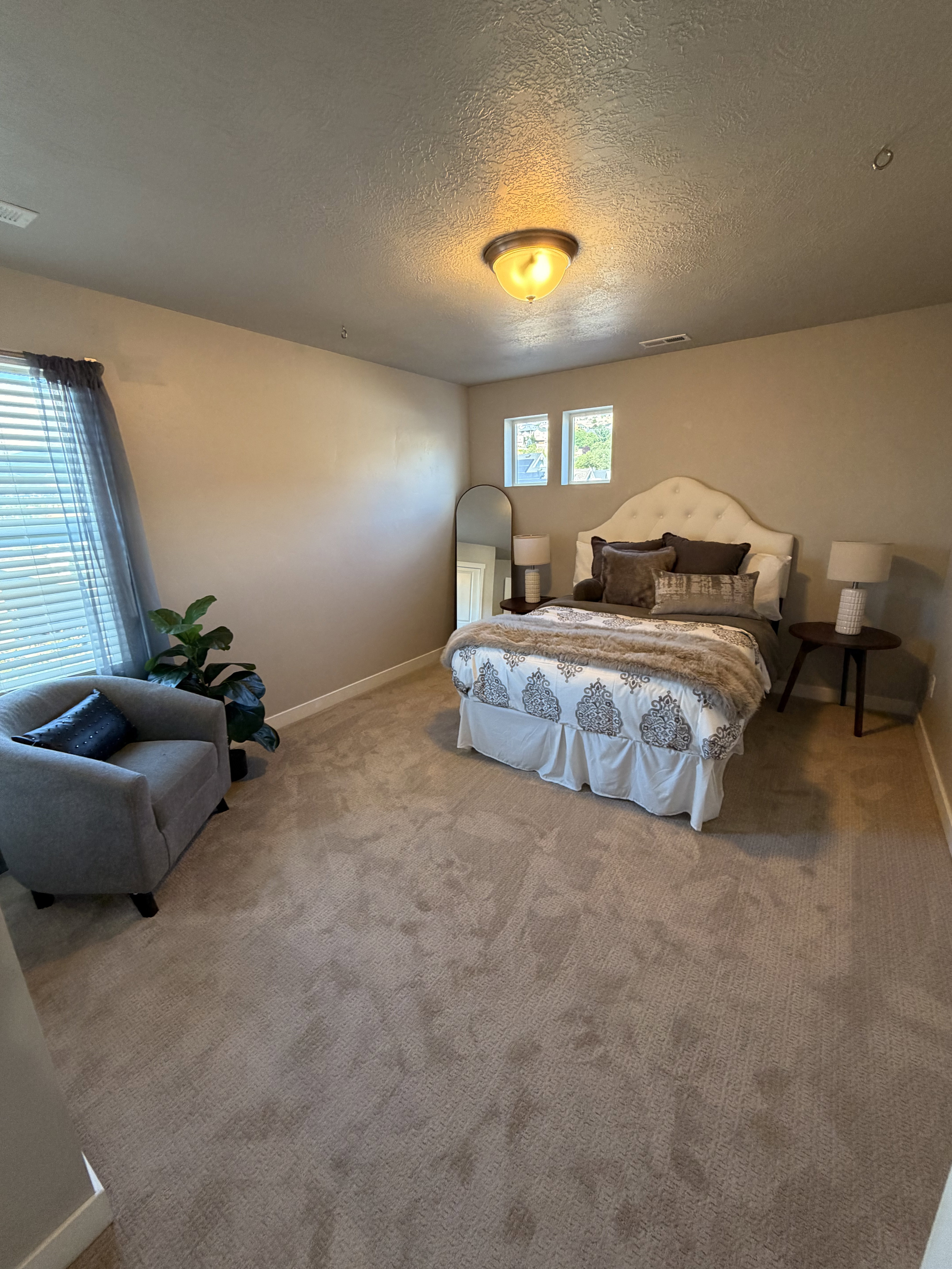 Carpeted room featuring a ceiling-mounted light fixture, two windows with blinds, a built-in upholstered headboard, and a neutral wall color