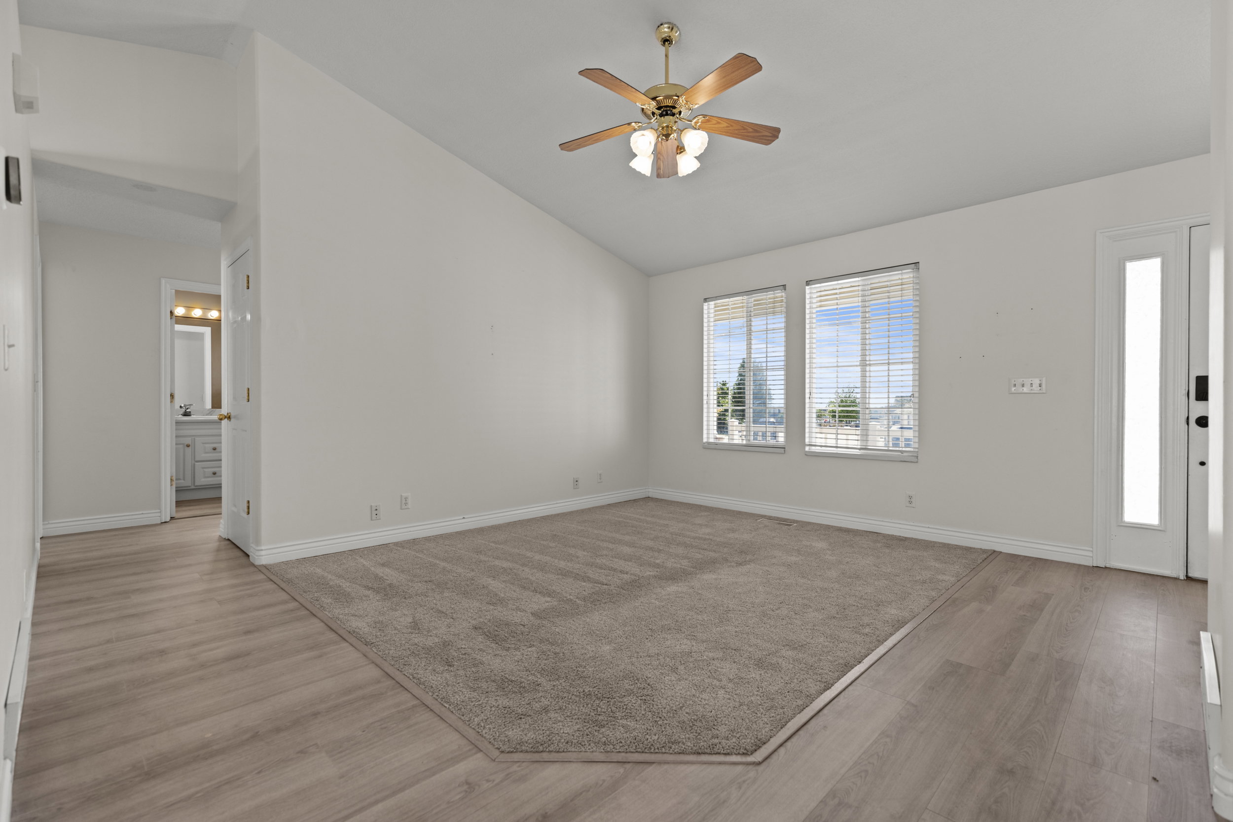 Vaulted ceiling room featuring a ceiling fan with light fixture, dual windows with blinds, a white paneled door with a full-length frosted glass insert, and a combination of light wood-finish flooring and patterned carpet