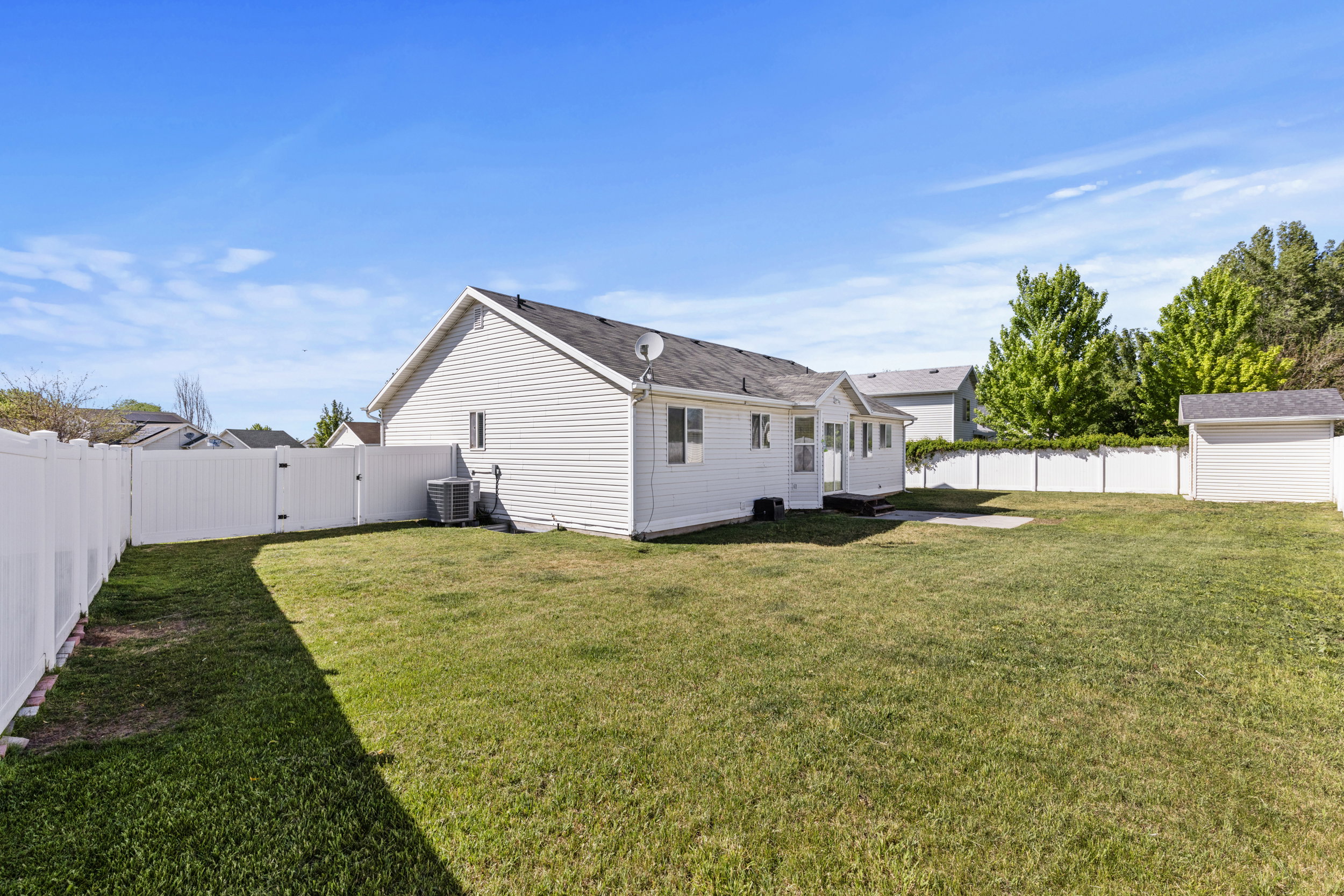 Expansive backyard featuring a manicured lawn, full white privacy fencing, concrete patio, and separate storage shed
