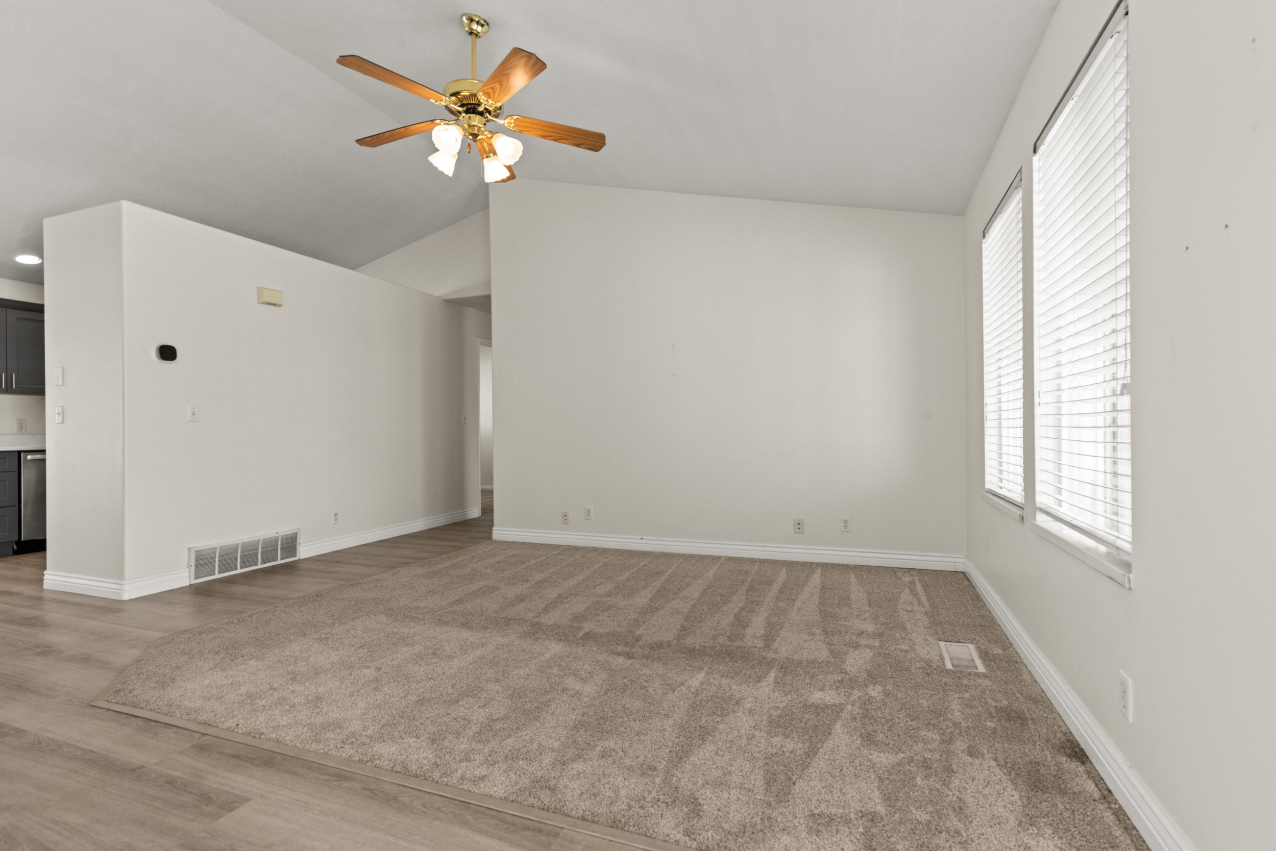 Carpeted living area featuring vaulted ceilings, two large windows with blinds, a ceiling fan with integrated lighting, and wood-finish flooring extending into an adjacent space