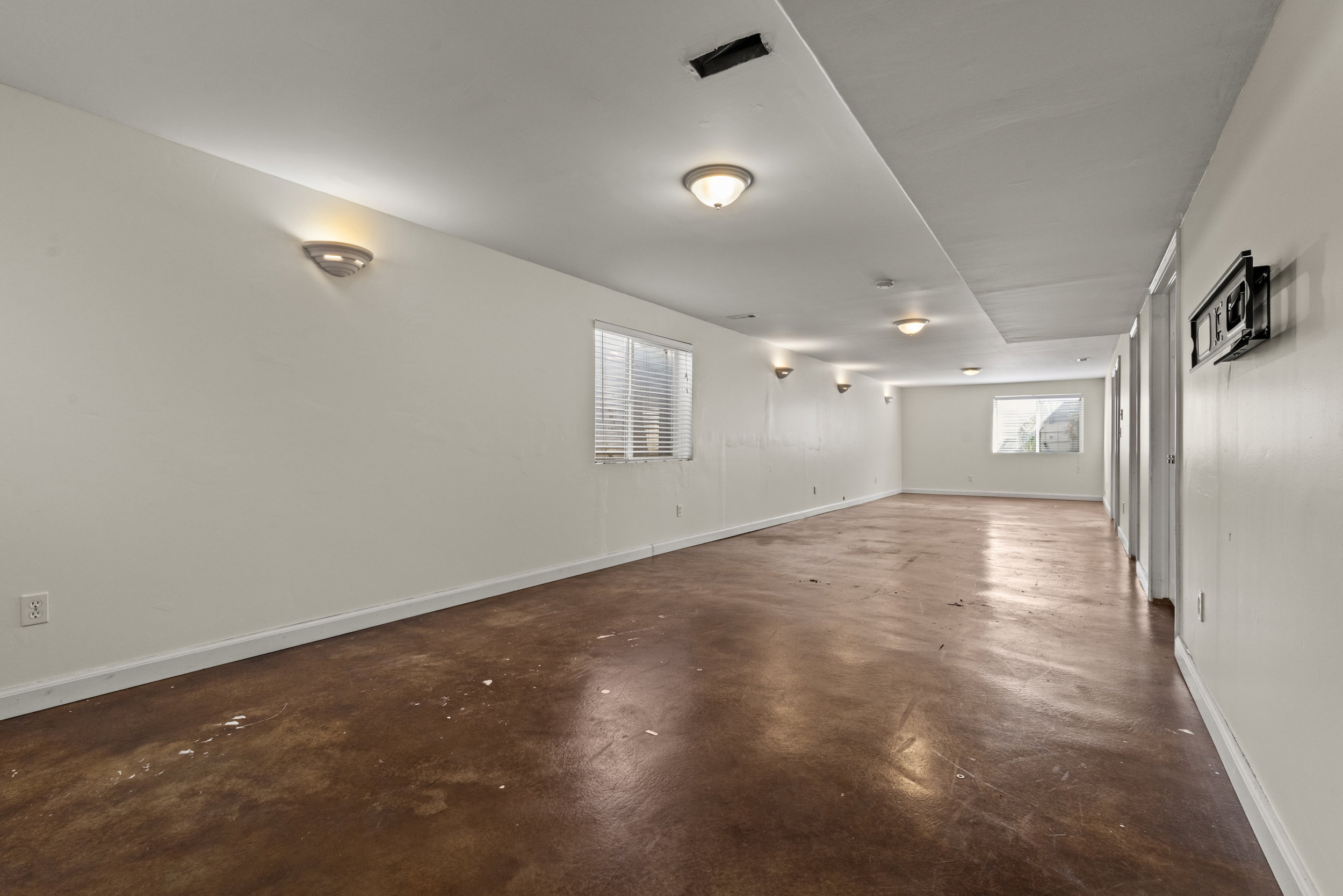 Spacious interior room featuring light-colored walls, multiple windows with blinds, built-in sconce lighting, and a durable brown flooring finish