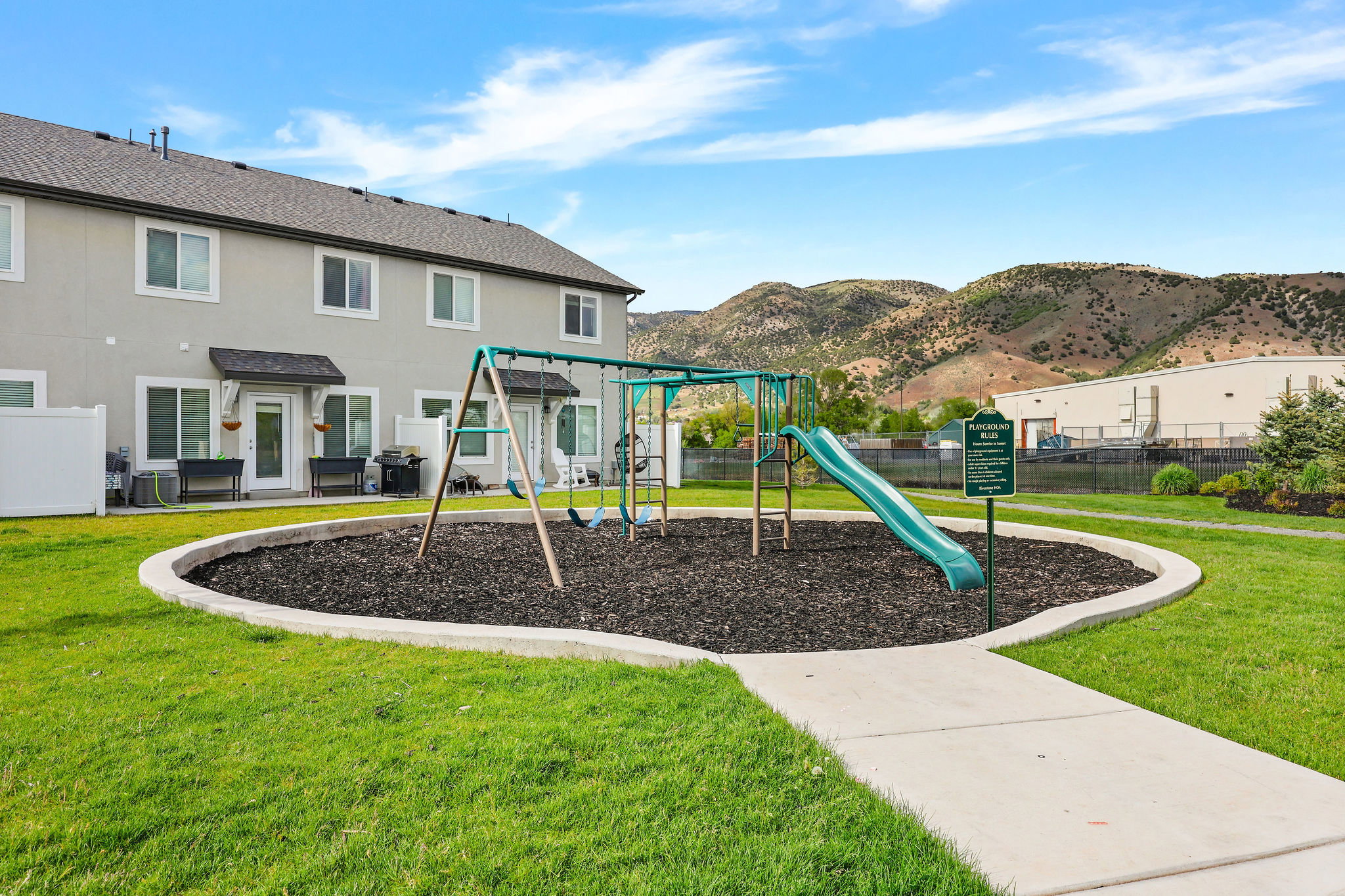Community playground featuring a swing set and slide on a mulch ground cover, surrounded by manicured green space