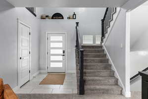 Entryway featuring a modern frosted-panel front door with black hardware, a contrasting gray tiled floor, and a carpeted staircase with dark wood-finish newel posts and spindles