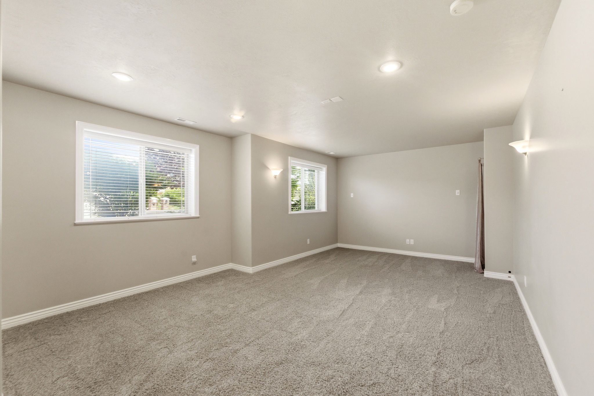 Spacious room featuring neutral carpeting, recessed ceiling lighting, two windows with blinds, and light-toned wall sconces