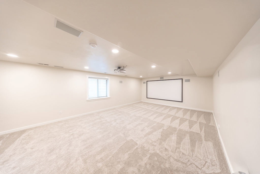 Expansive media room featuring a large projection screen, mounted projector, recessed lighting, patterned carpet, and a window with blinds