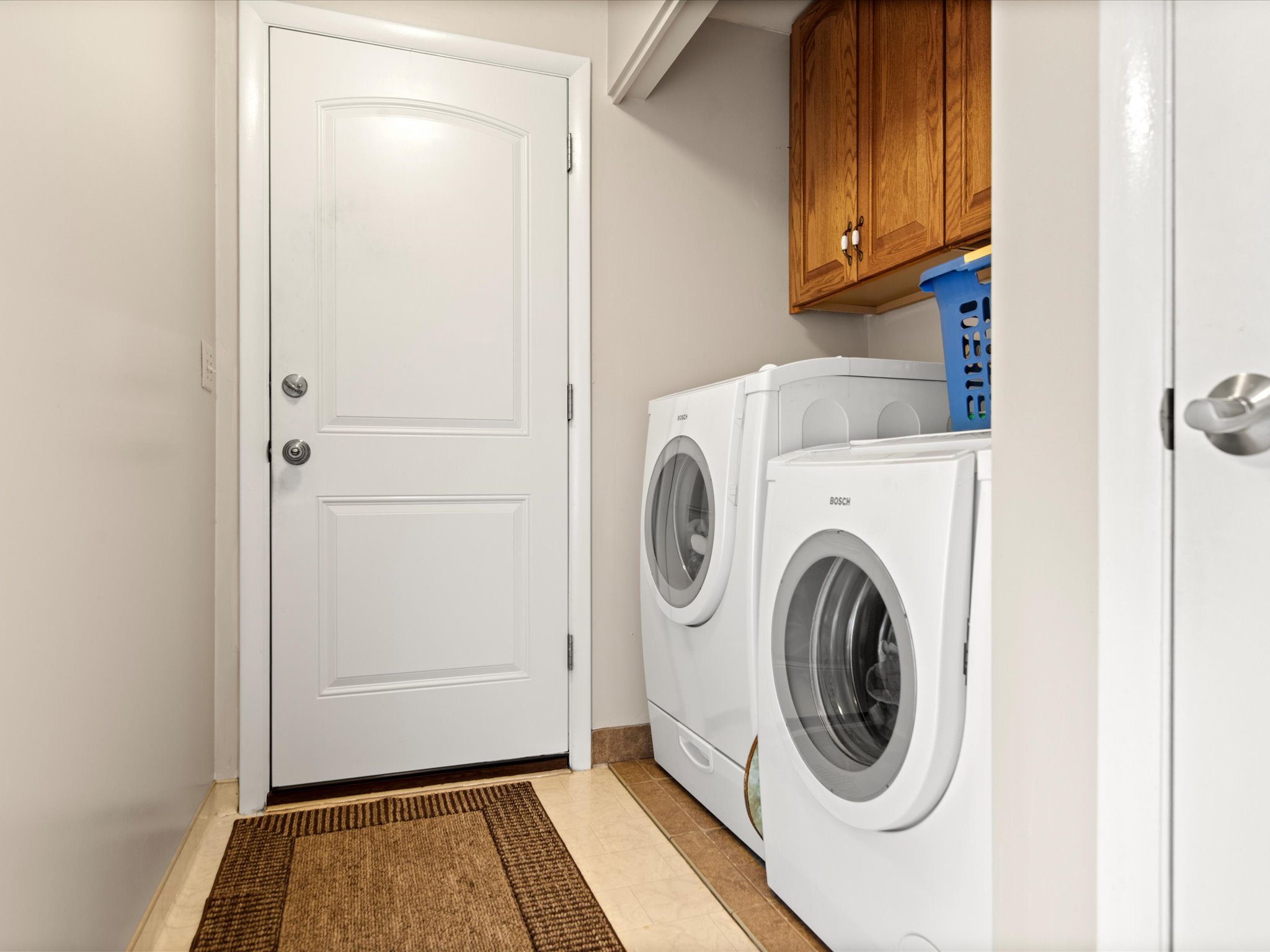 Dedicated laundry area featuring a white paneled door, light-toned wall paint, natural wood-finish cabinetry, and neutral-toned floor tiling