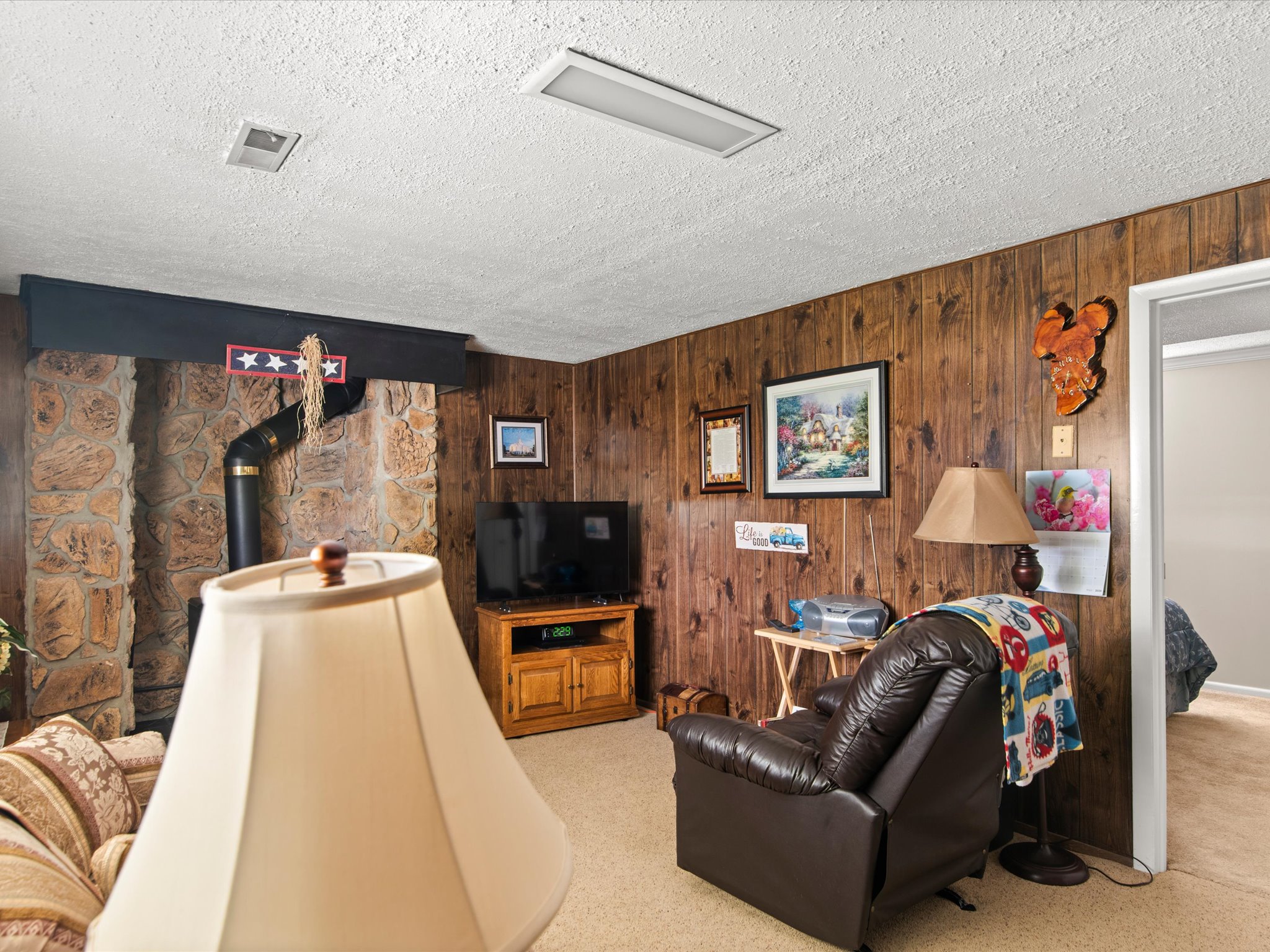 Stone-clad fireplace with black stovepipe, wood-finish wall paneling, and neutral carpet flooring