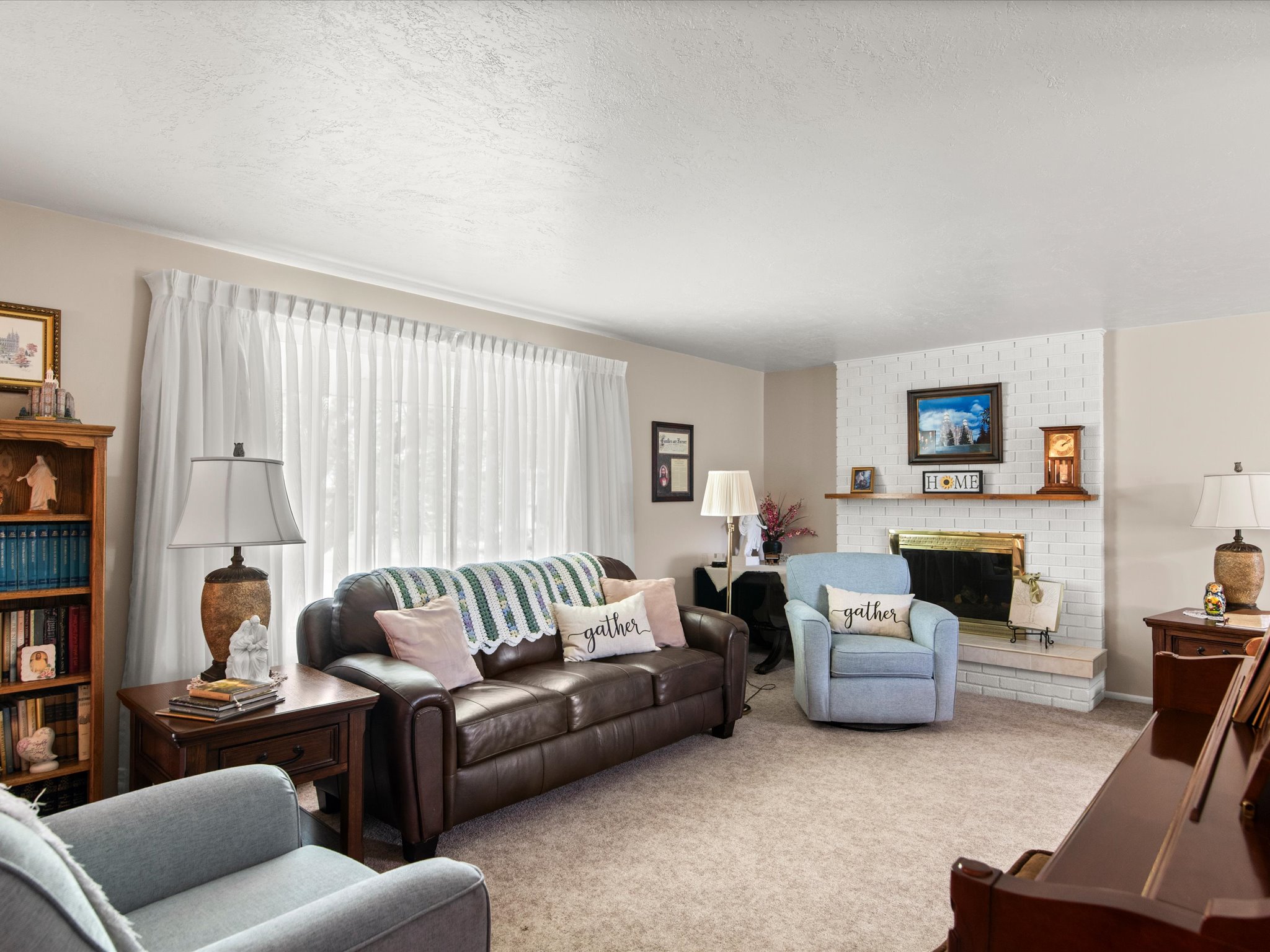 Carpeted living space featuring a white brick fireplace with a wood mantle, a large window with sheer white curtains, and neutral wall paint