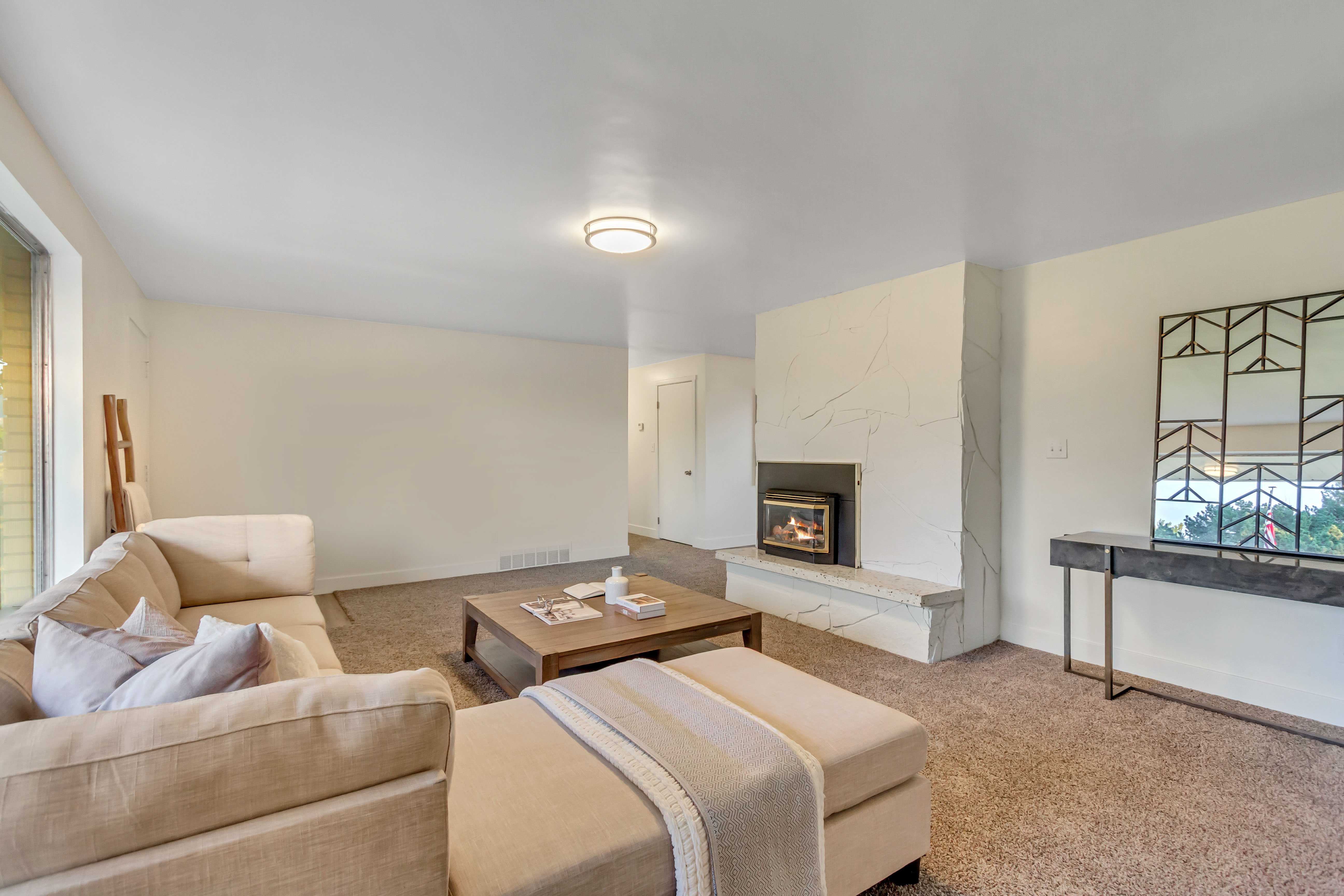 Spacious living area featuring a prominent fireplace with a textured surround and hearth, neutral-toned carpeting, and a large window providing natural light