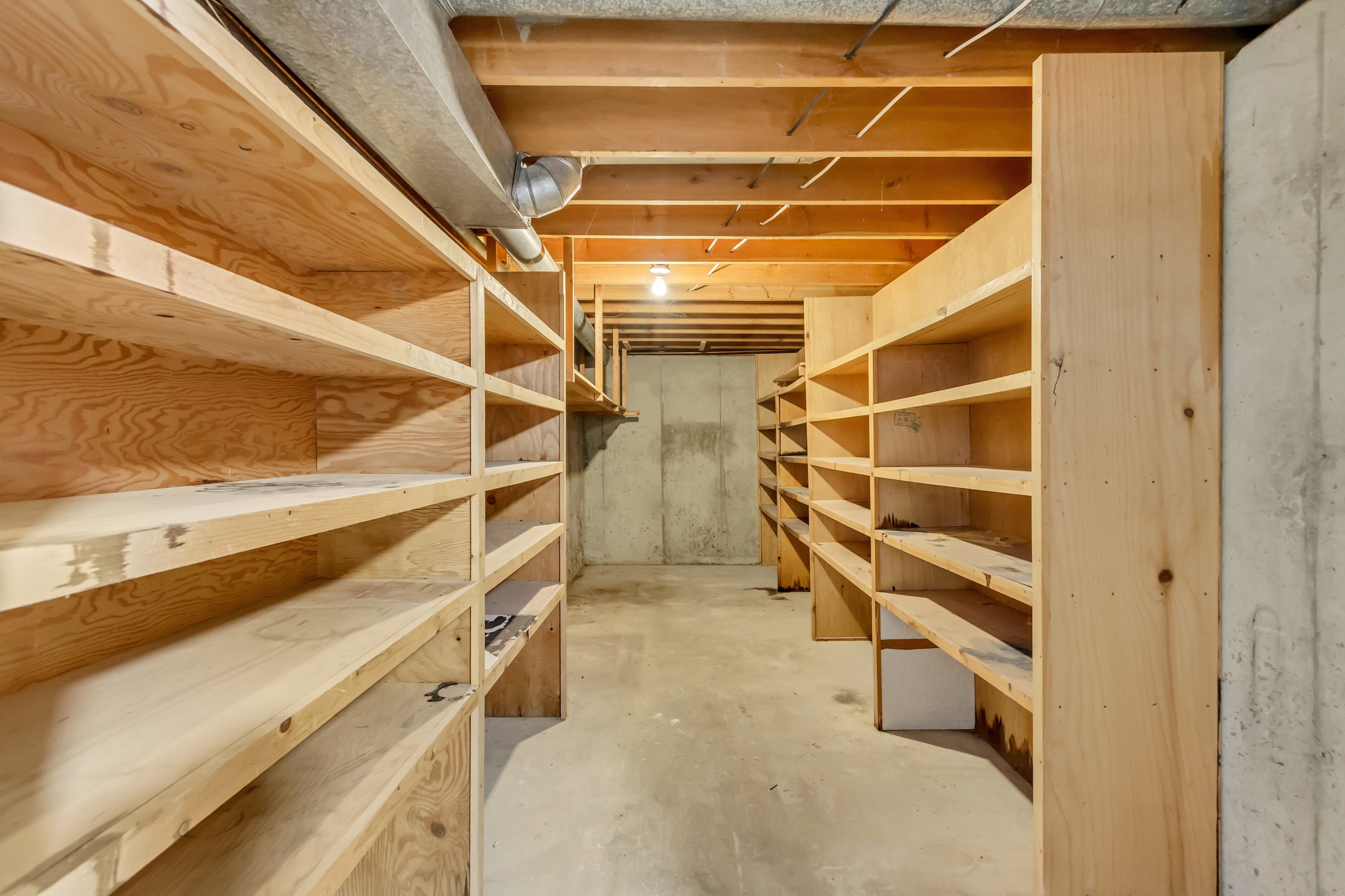 Basement storage area featuring extensive built-in wooden shelving, concrete walls, exposed joists, and concrete flooring