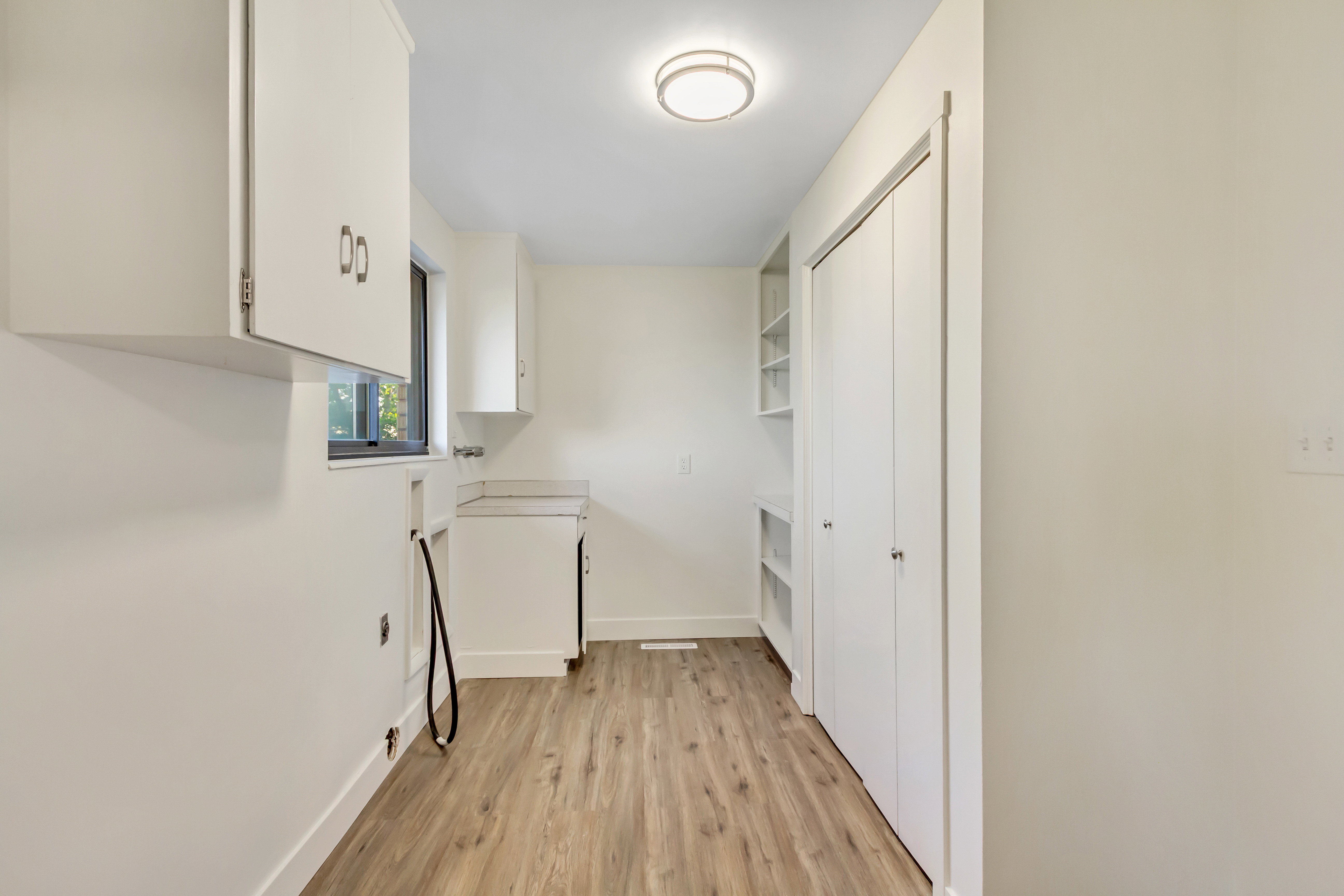 Utility room featuring wood-finish flooring, white cabinetry, a built-in countertop, a window, and a bifold door closet with integrated shelving