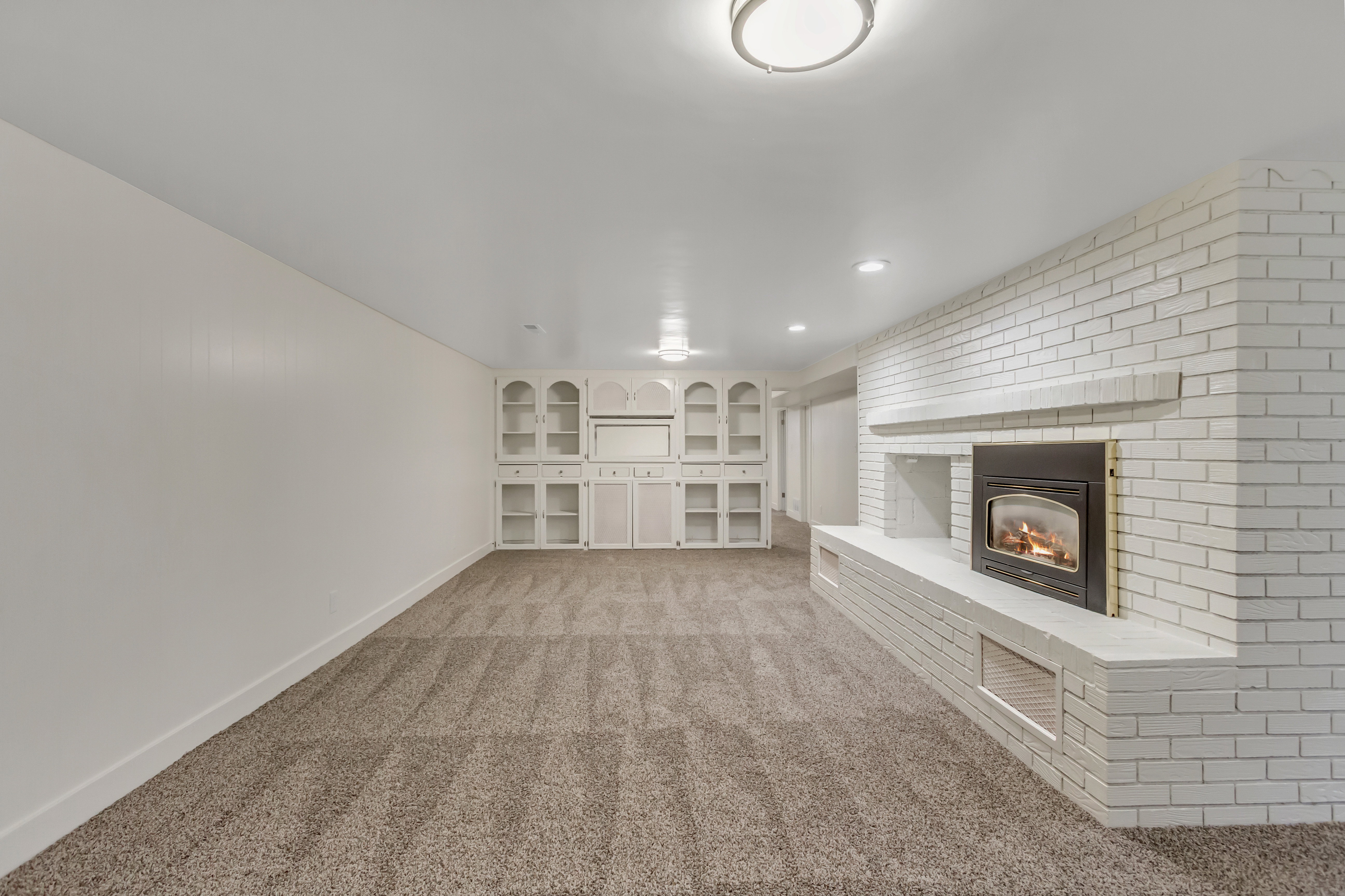 Spacious room featuring a white painted brick fireplace with a raised hearth, built-in shelving, recessed lighting, and neutral carpet