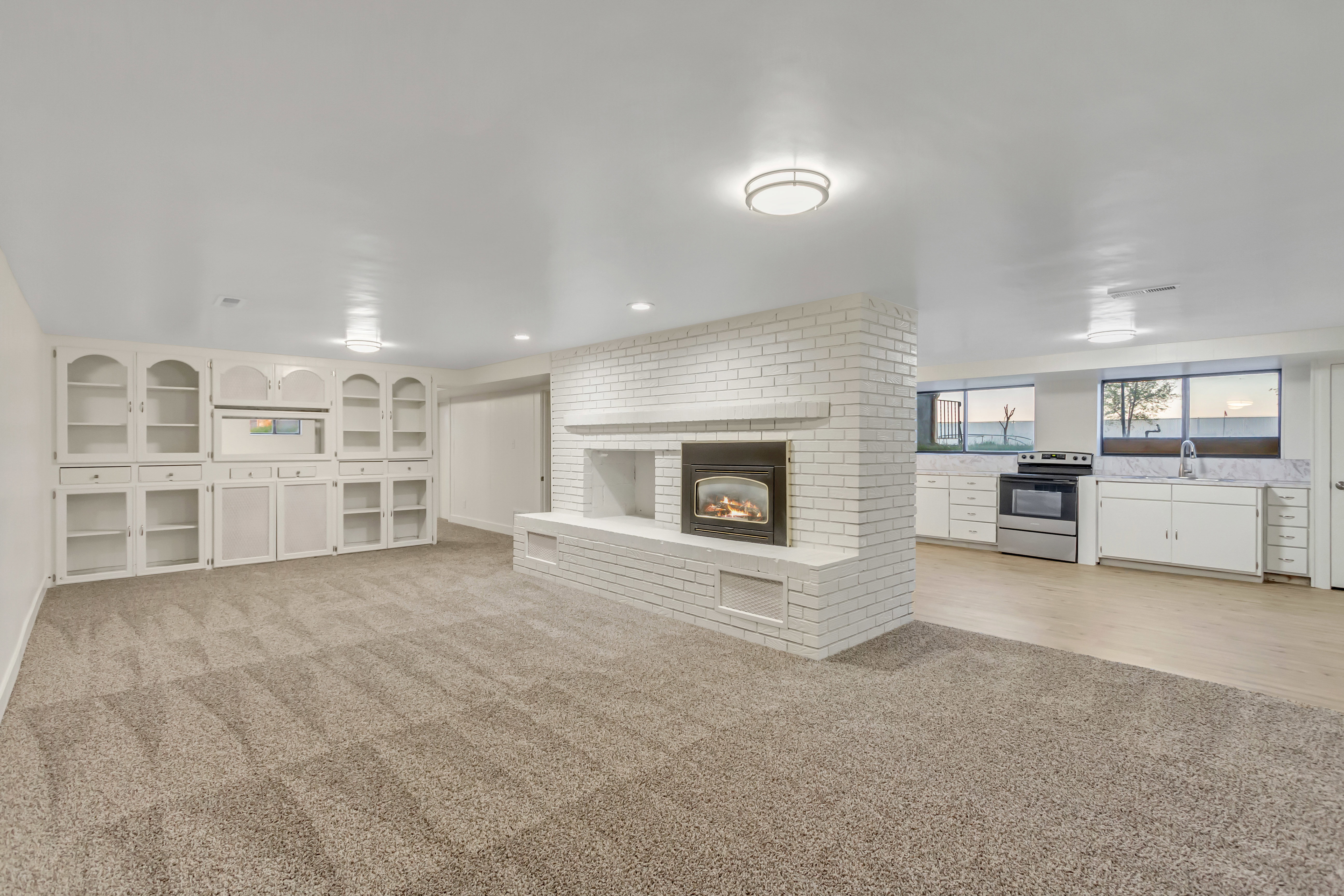 Living area with a white brick fireplace, built-in cabinetry, and neutral carpet, opening to a kitchen featuring light wood-finish flooring, white cabinetry, and stainless steel appliances