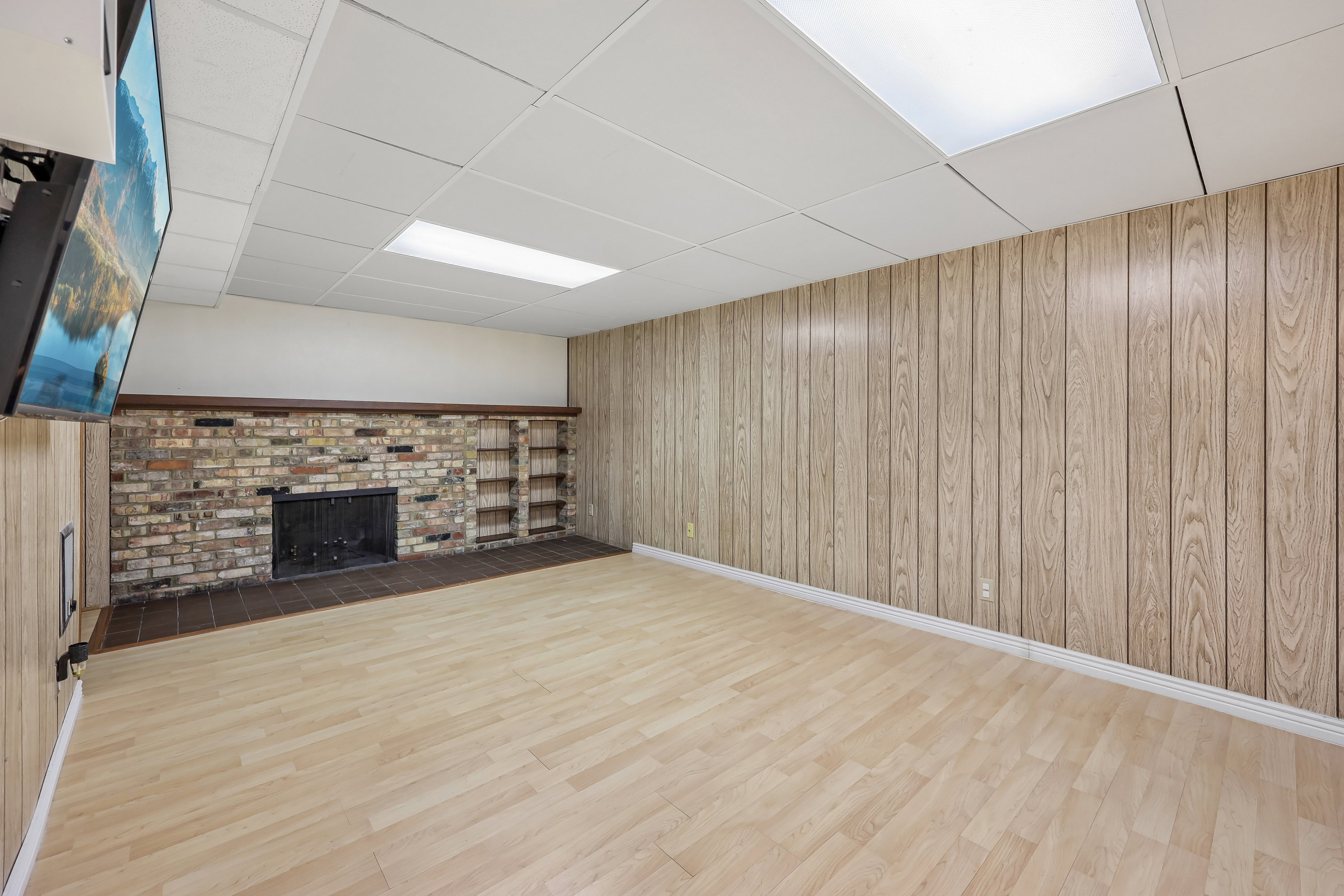 Spacious room featuring light wood-finish flooring, vertical wood paneling, a brick fireplace with hearth, and recessed ceiling lighting