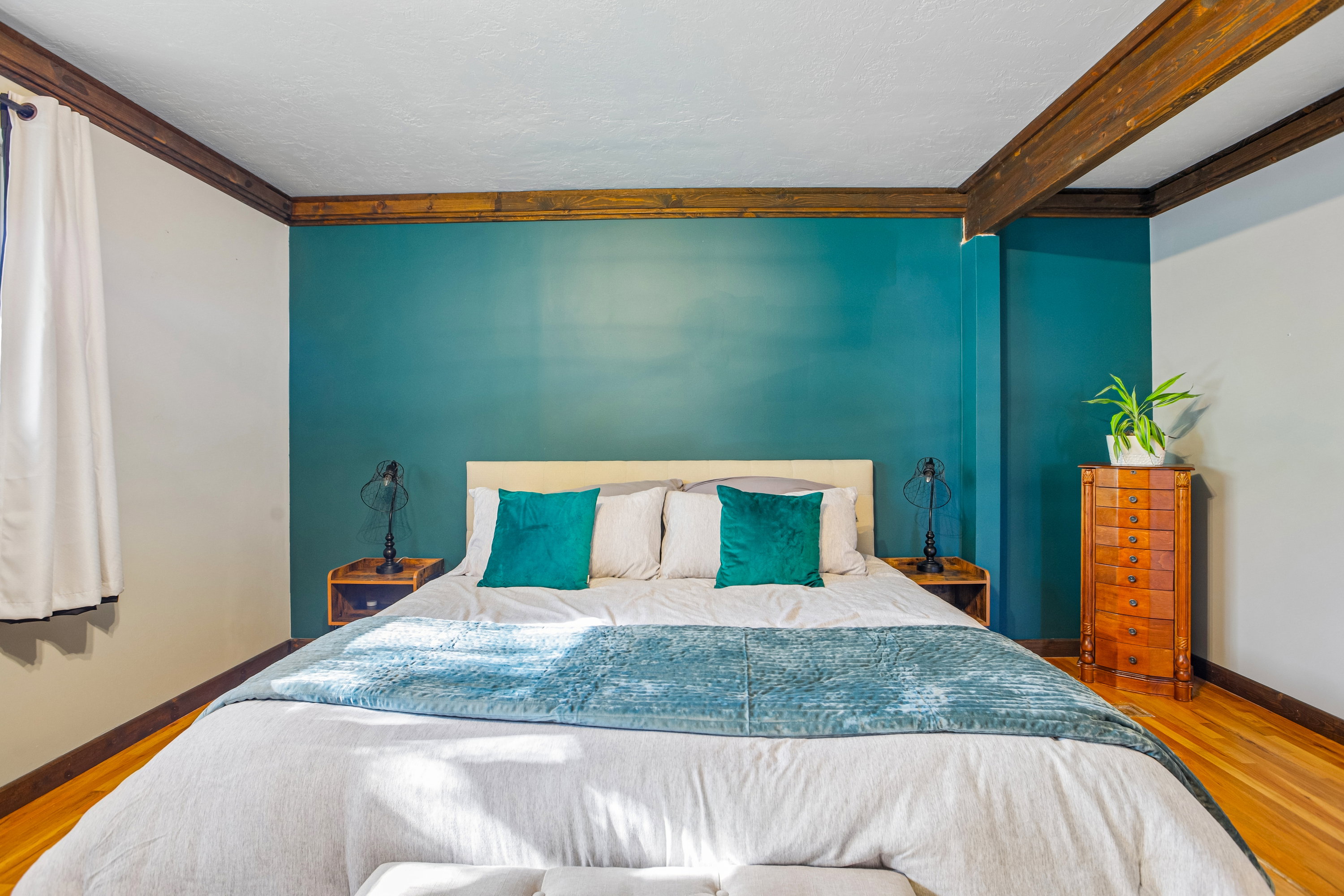 Bedroom featuring a teal accent wall, wood-finish flooring, exposed ceiling beams, and a window with drapes