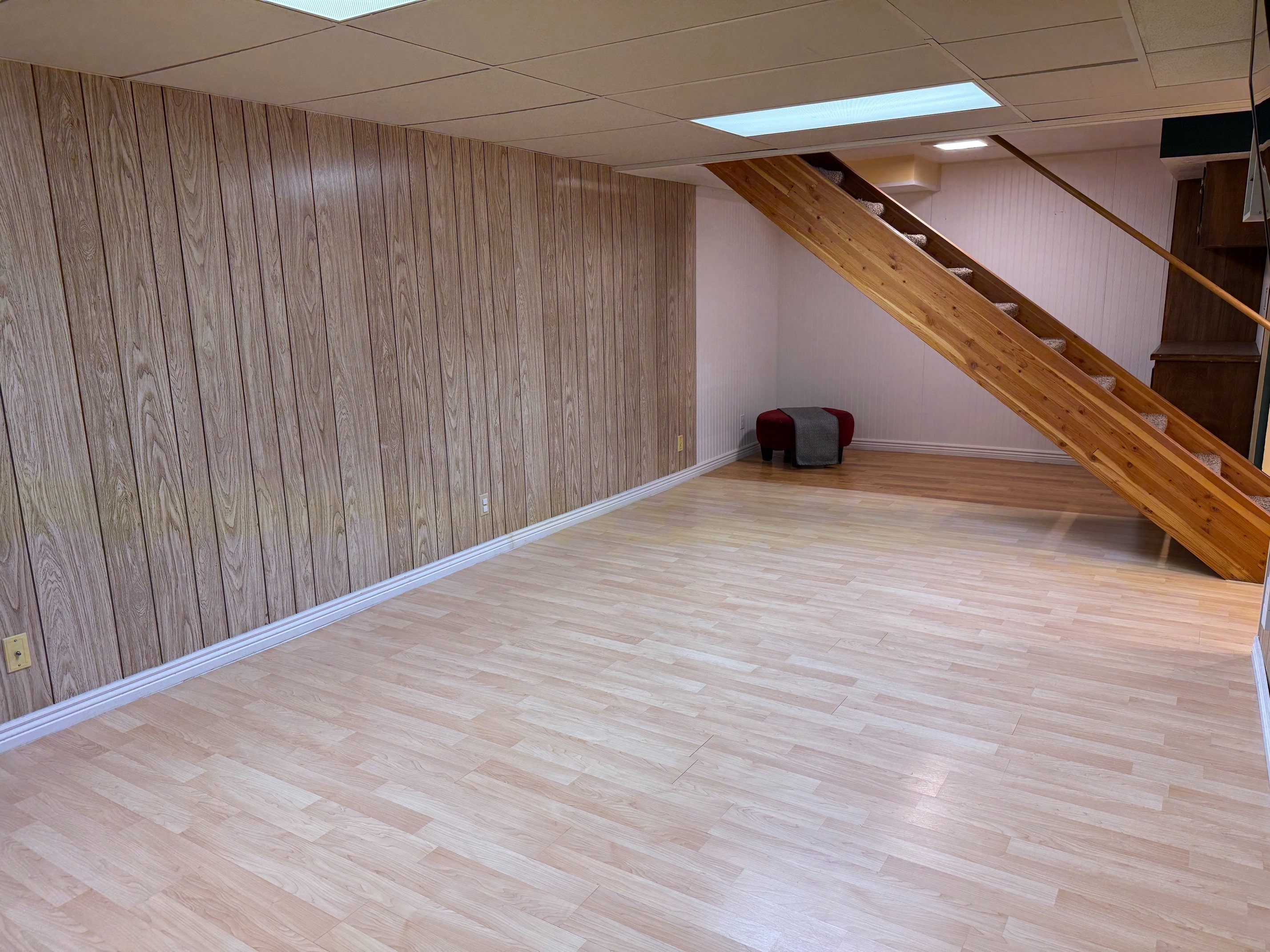 Spacious open room featuring wood-finish flooring, accent wall paneling, white baseboards, and a natural wood staircase with carpeted treads