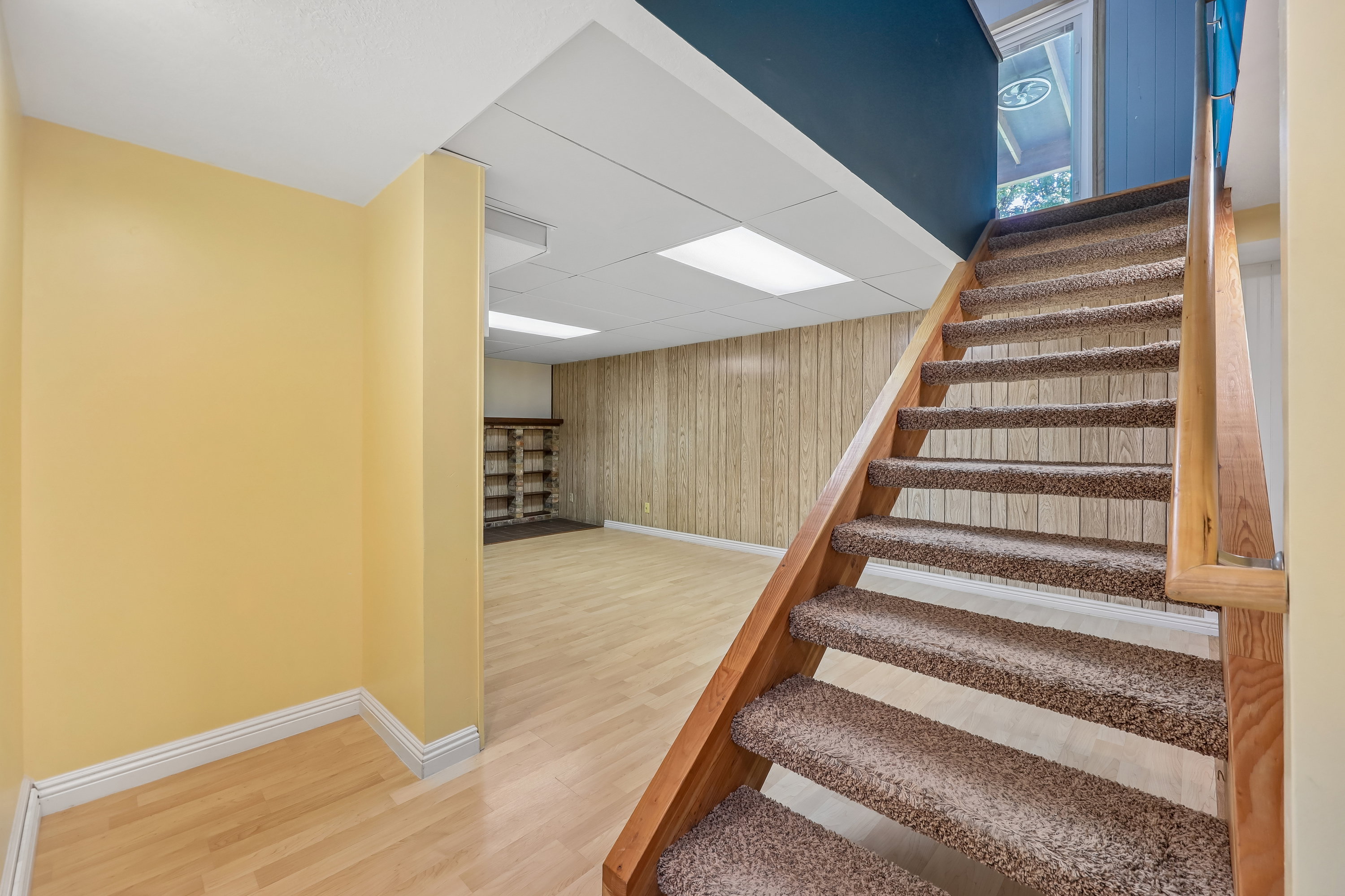 Carpeted staircase with wood handrail, leading to a finished lower level featuring wood-finish flooring, wood paneling, and a built-in shelving unit