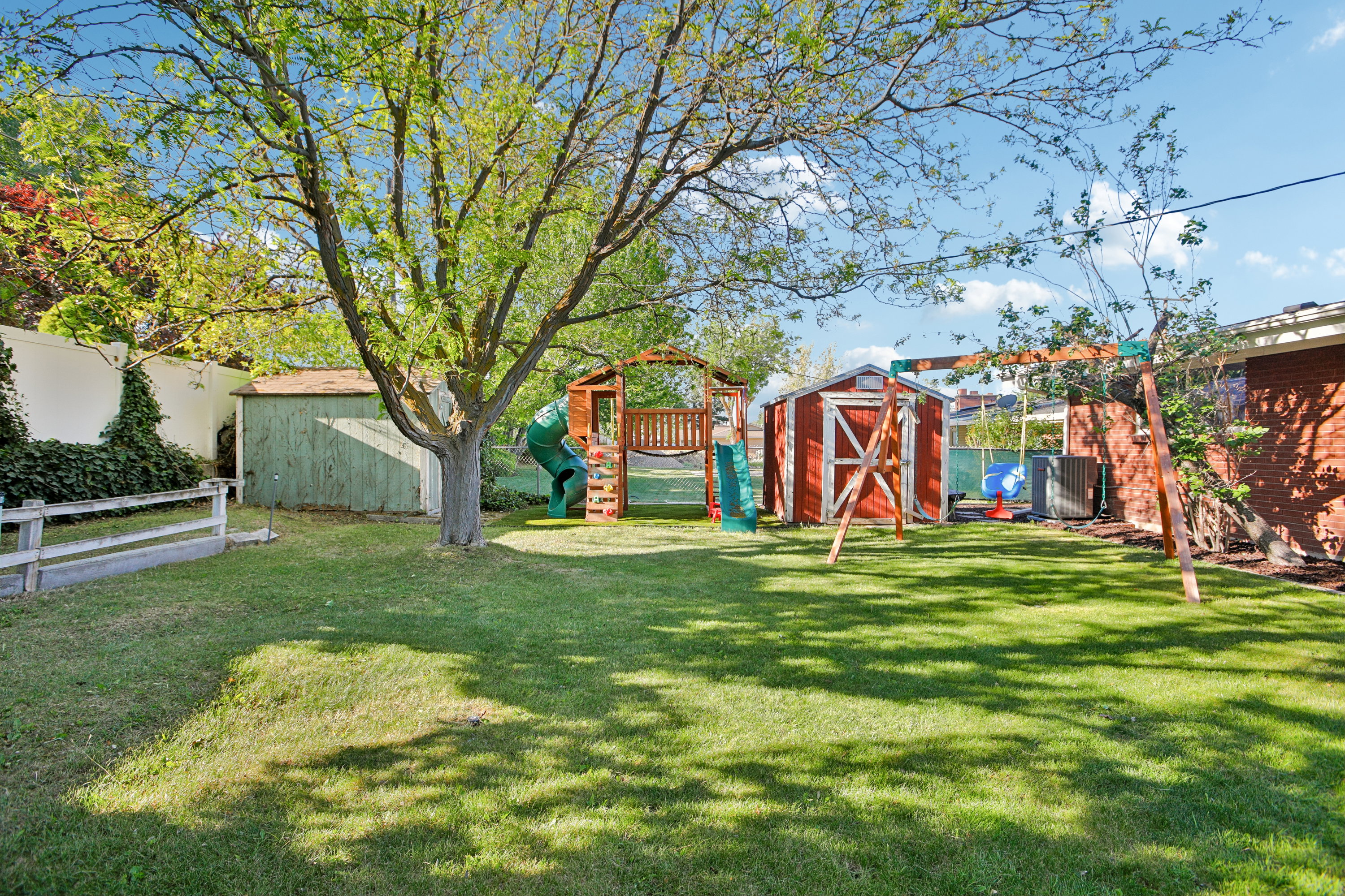 Expansive grassy yard featuring mature shade trees, a red storage shed, a green shed, and a privacy fence