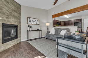 Living area featuring a floor-to-ceiling tiled fireplace, wood-finish flooring, a ceiling fan, and a pass-through opening to the kitchen with a wood beam accent