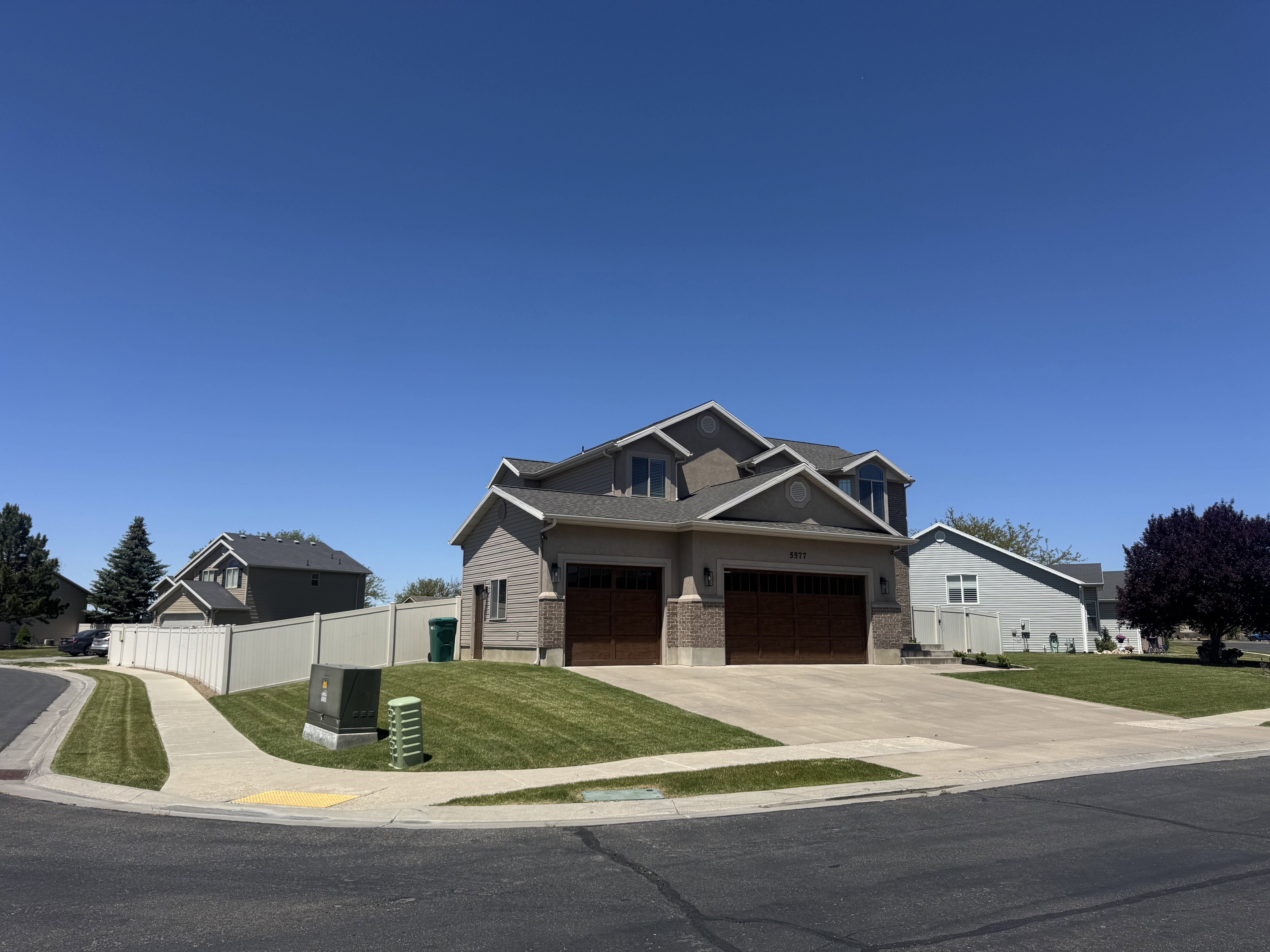 Two-story residence featuring a tan siding exterior with brick accents, prominent dormer windows, a two-car garage with wood-finish doors, and a concrete driveway