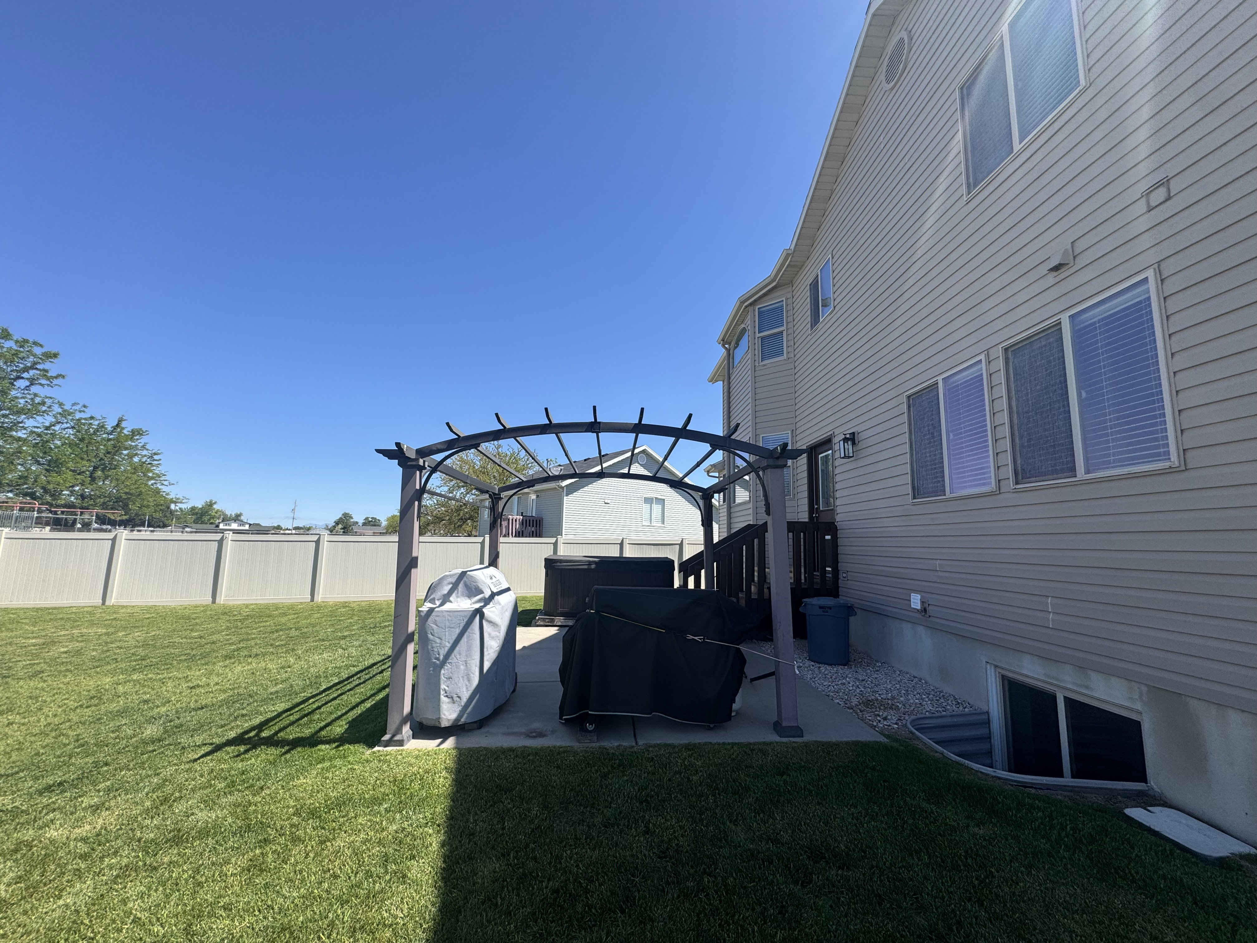 Spacious backyard featuring a manicured lawn, a concrete patio with a dark metal pergola, and a visible basement window well